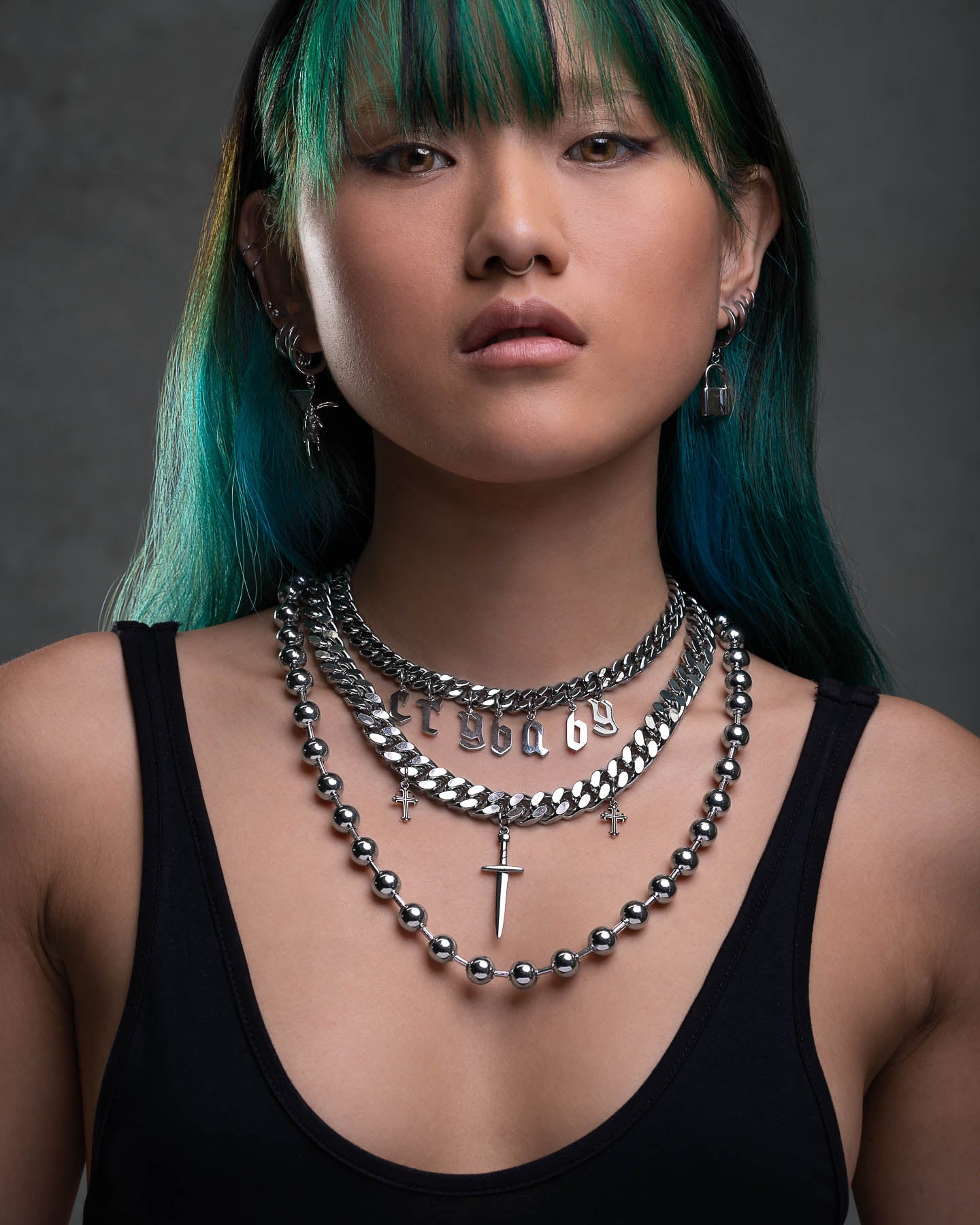 A person with green and black hair wears the Crybaby Stack—multiple silver necklaces including a Crybaby necklace and cross pendant—over a black tank top, paired with hoop and dangle earrings, against a gray background.