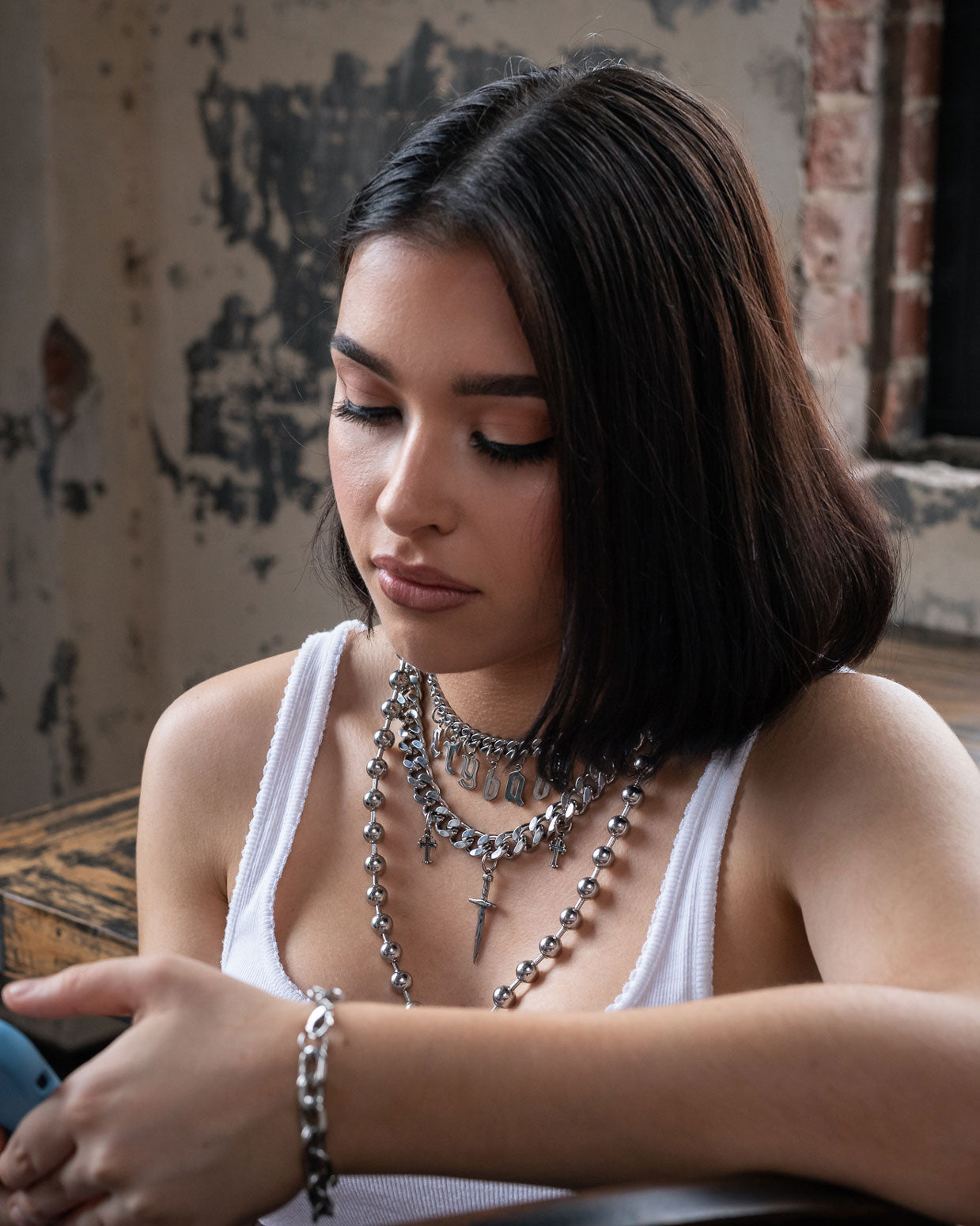 A young woman with straight dark hair wears the Crybaby Stack festival jewelry, including a small choker, and a white tank top indoors. She looks down at her phone with a neutral expression against a rustic, worn wall background.