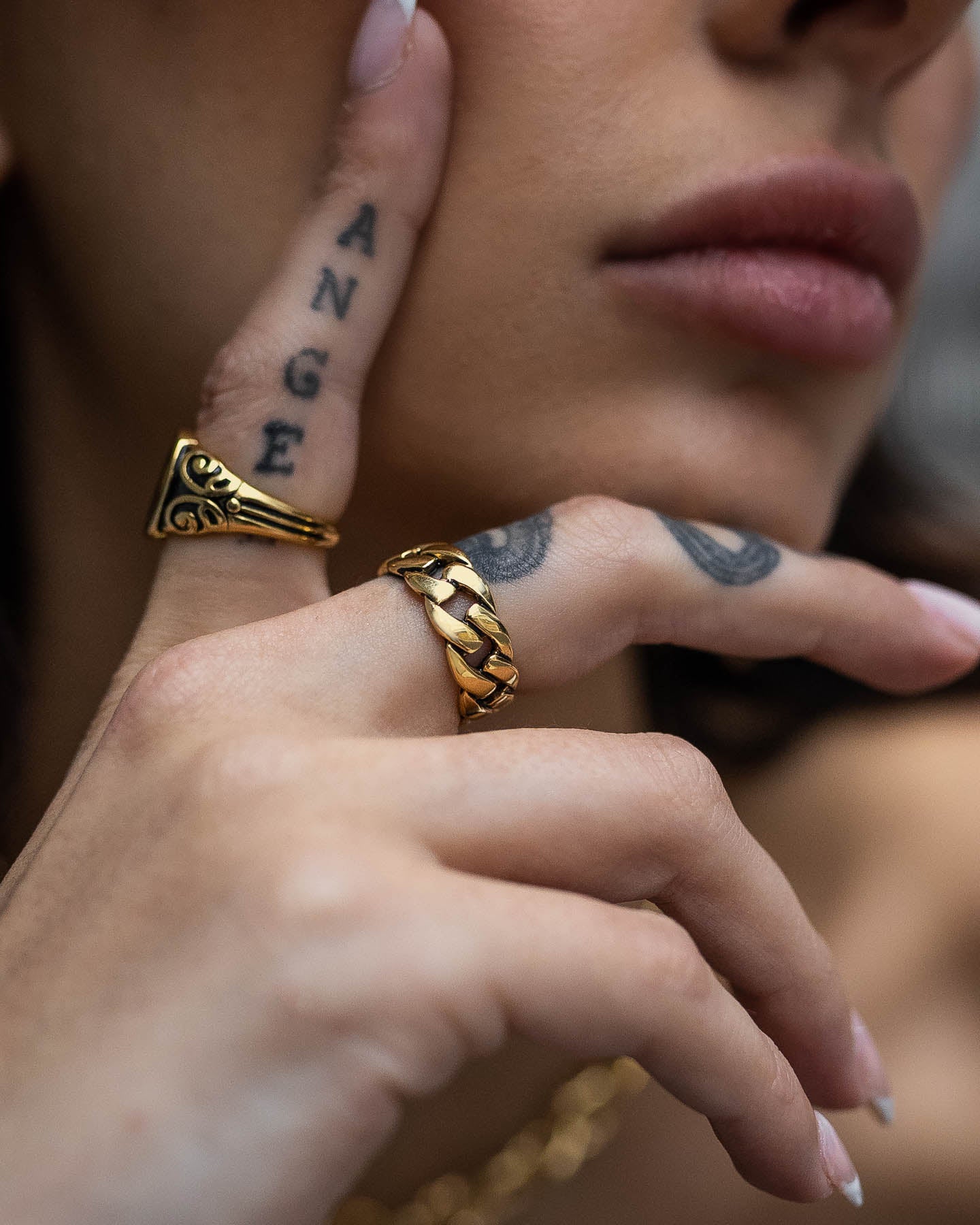 A close-up of a light-skinned person’s hand and face highlights the Cuban Link Ring (Gold) and other gold rings on their fingers, with “ANGE” tattooed across the index finger. The person has full lips.