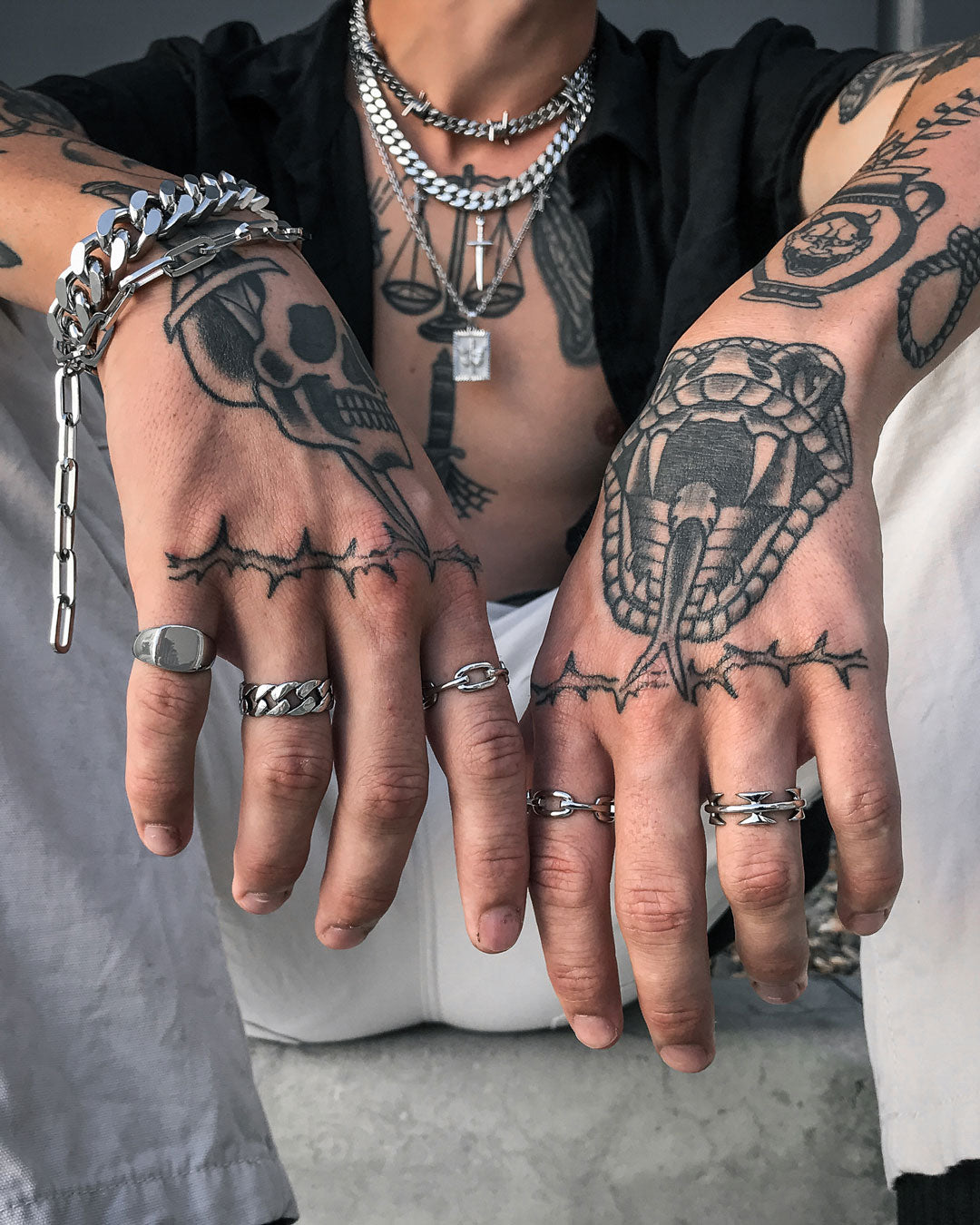 A person with snake and skull hand tattoos, wearing multiple silver bracelets, a men’s ring, the Cuban Link Ring, layered chain necklaces over a partly open black shirt and white pants.
