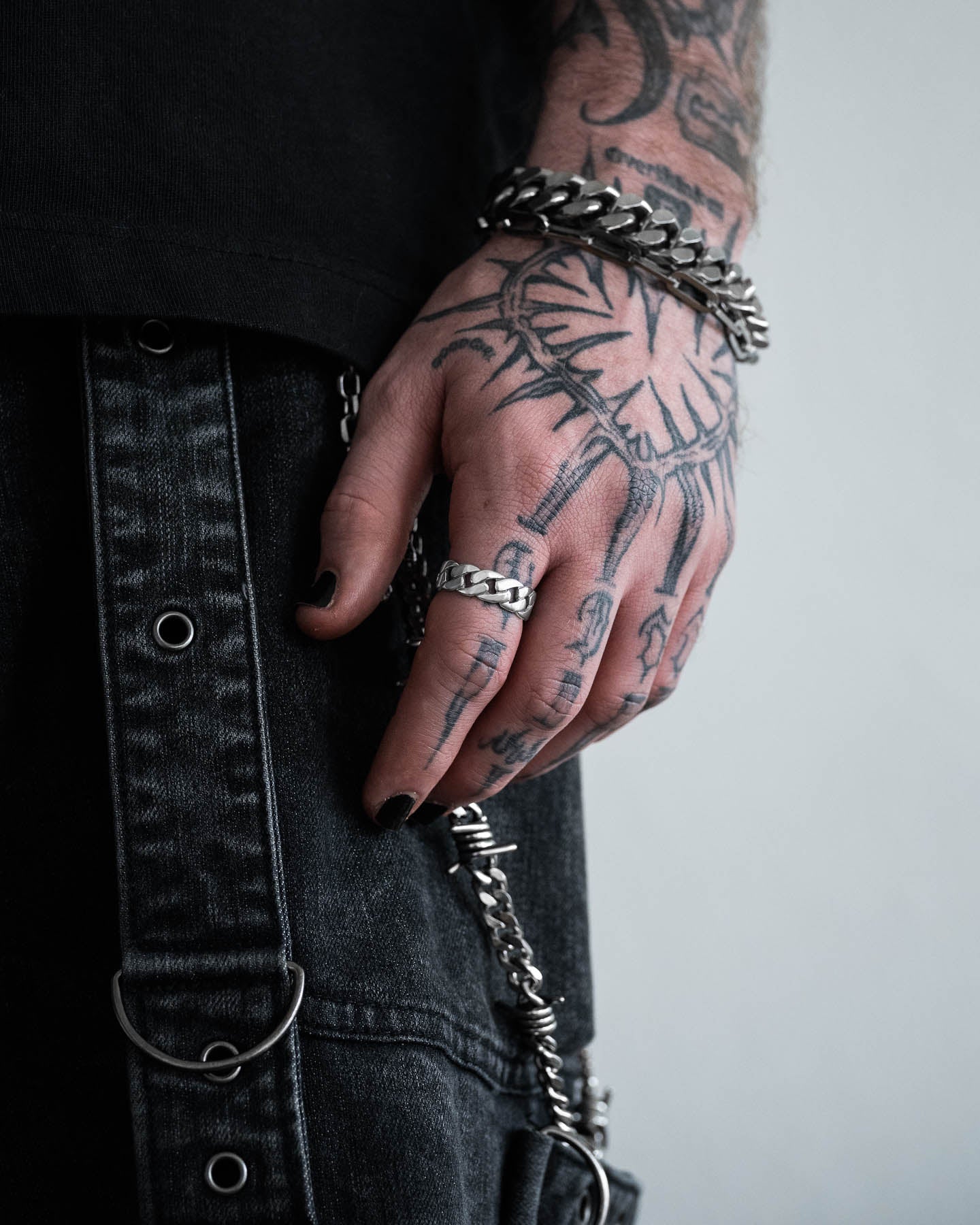 A tattooed hand with black nail polish wears the Cuban Link Ring and silver rings, along with a chain bracelet, resting beside black denim pants featuring metal eyelets and chain accessories.