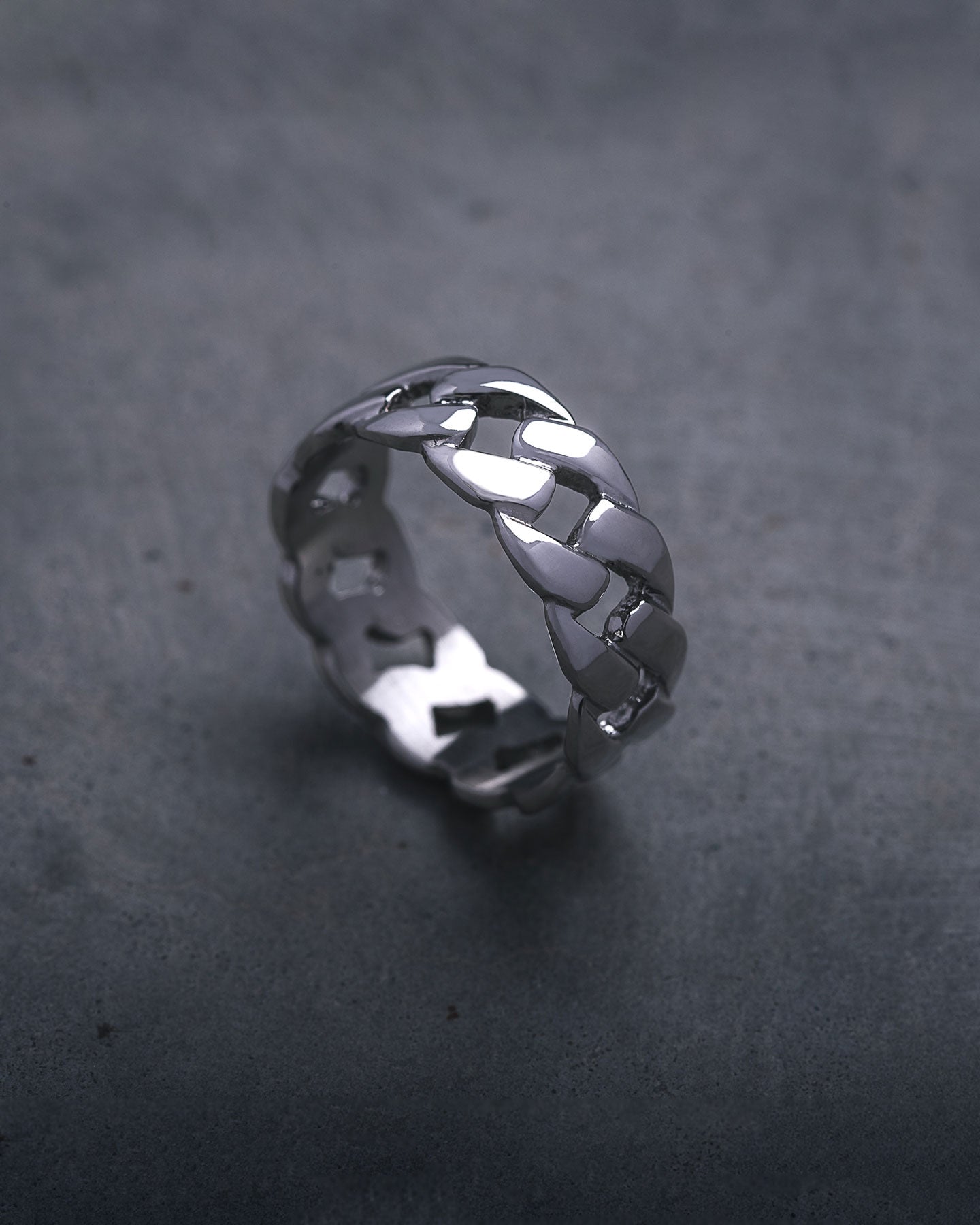The Cuban Link Ring, featuring a chunky silver interlocking chain design, is displayed upright on a dark, textured surface.