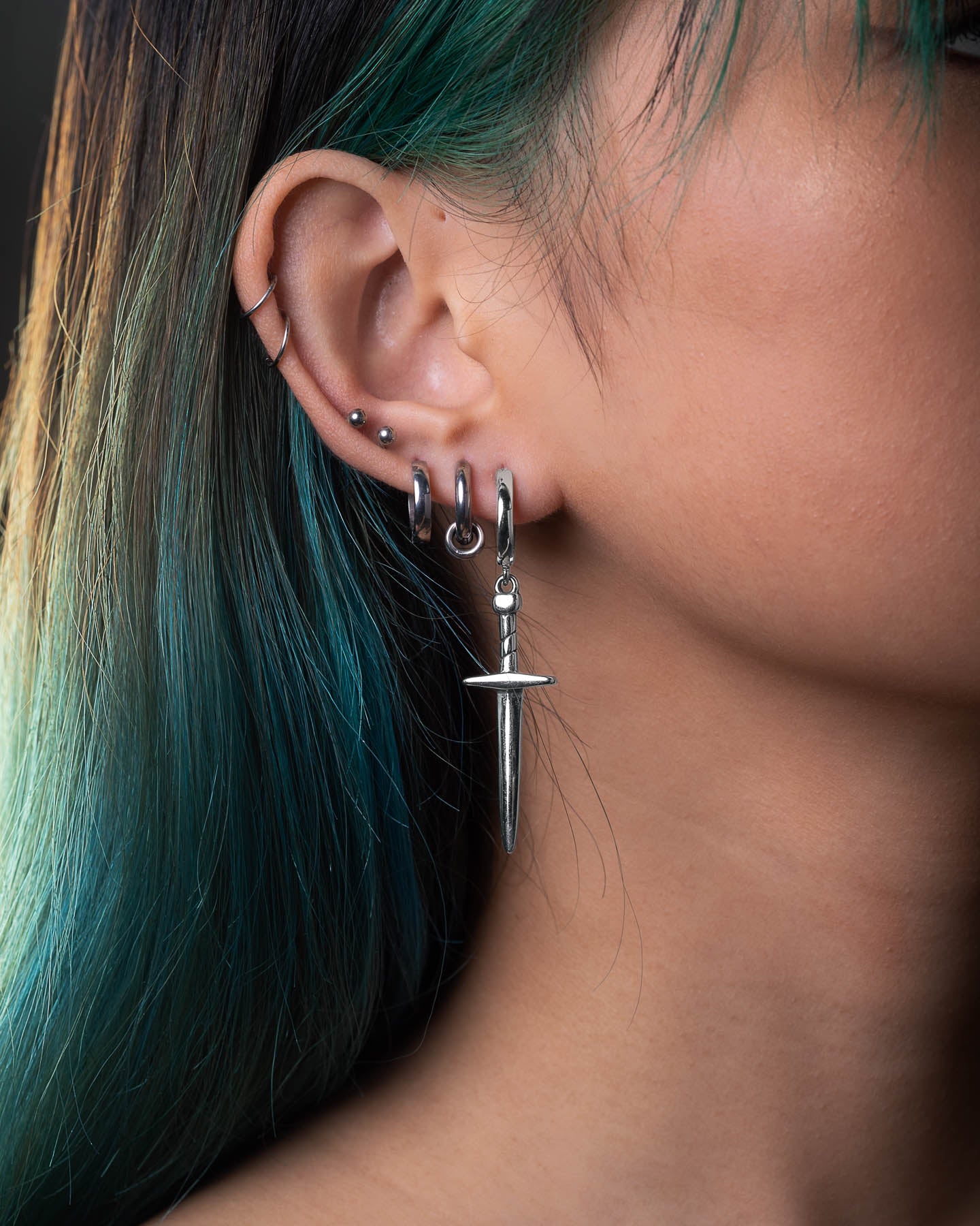 A close-up shows a person with teal and black hair wearing multiple silver hoops and the bold Dagger Earring, creating a striking statement look.