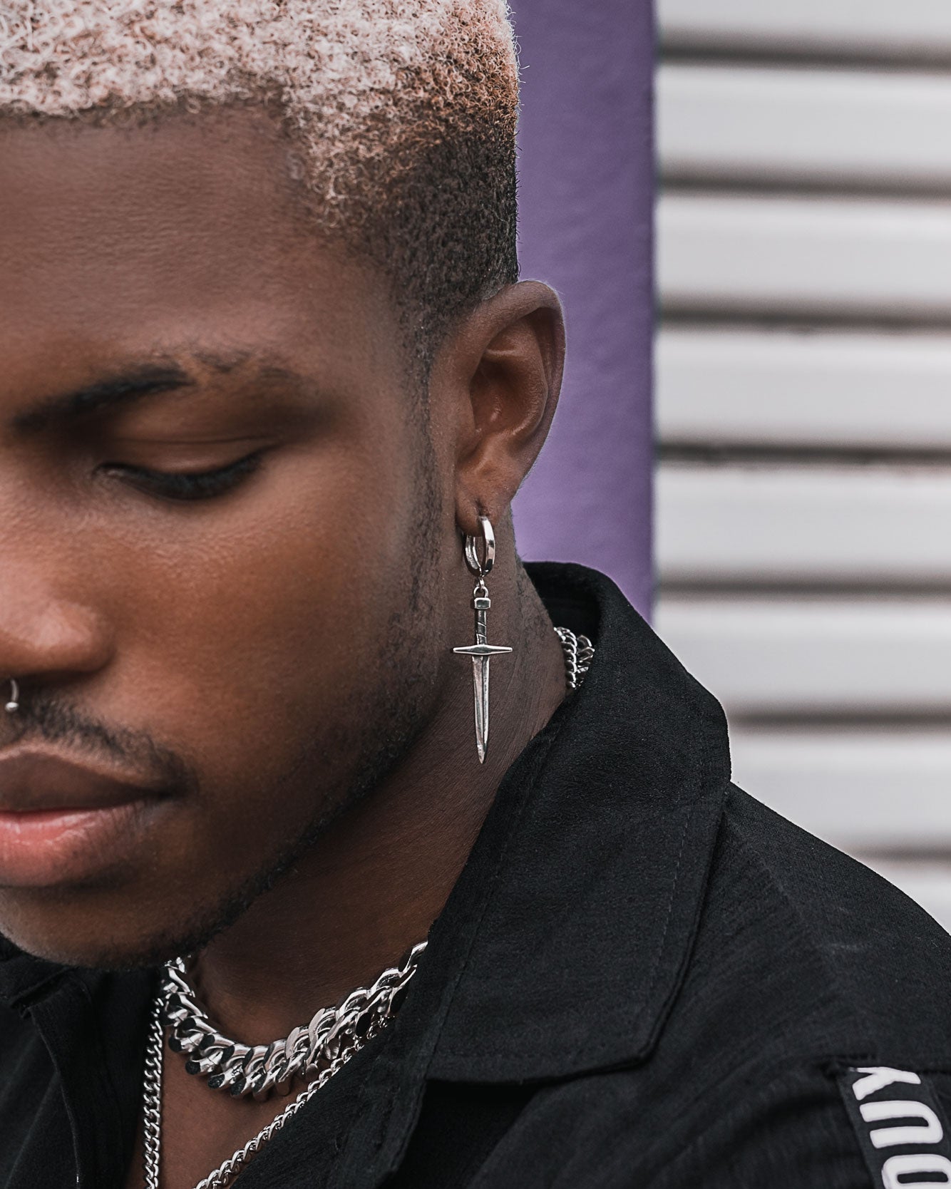A person with short, bleached hair wears the Dagger Earring, a silver nose ring, and layered chain necklaces as statement jewelry with a black shirt, standing before a striped background.