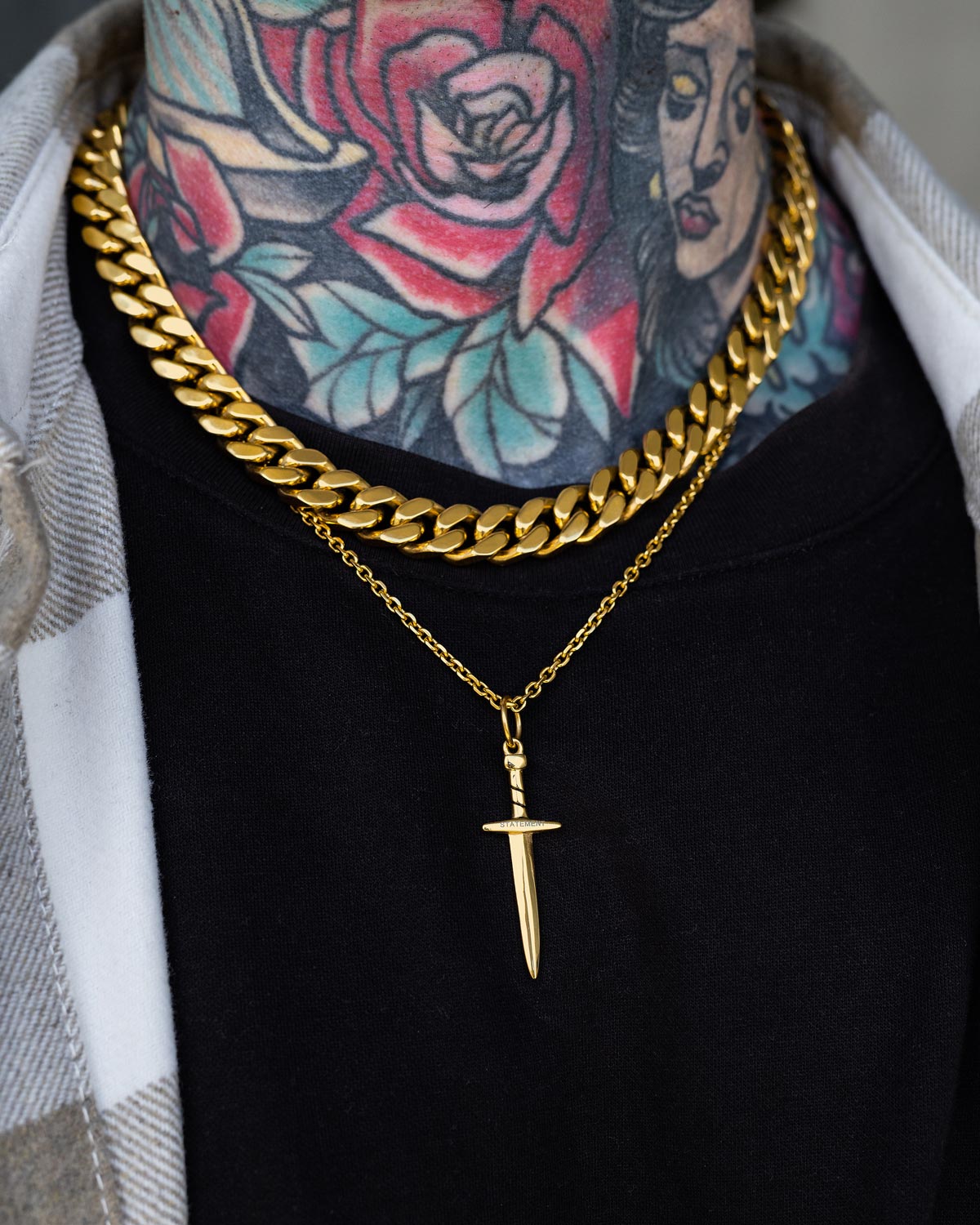 A close-up of a person's neck wearing Statement Collective's Dagger Pendant (Gold) on two gold chains—one thick, one thin. Colorful rose and woman's face tattoos pop against a black shirt and plaid jacket.