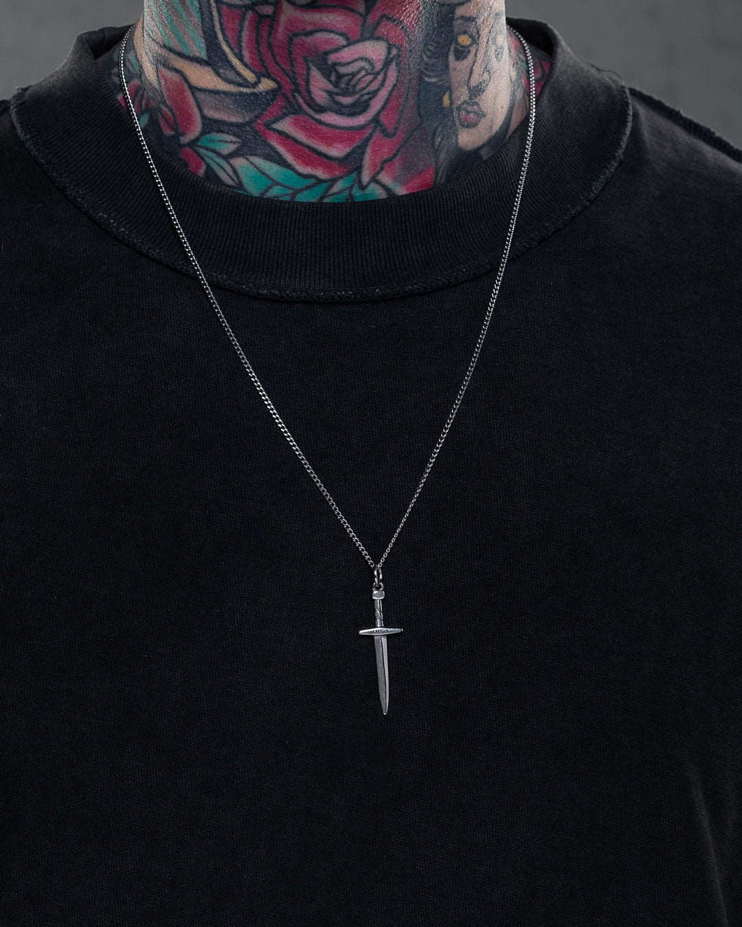 A person wears a black shirt and the Dagger Pendant necklace. Their neck and lower face are covered in colorful tattoos, like a rose and a woman's face. The background is neutral and blurred.