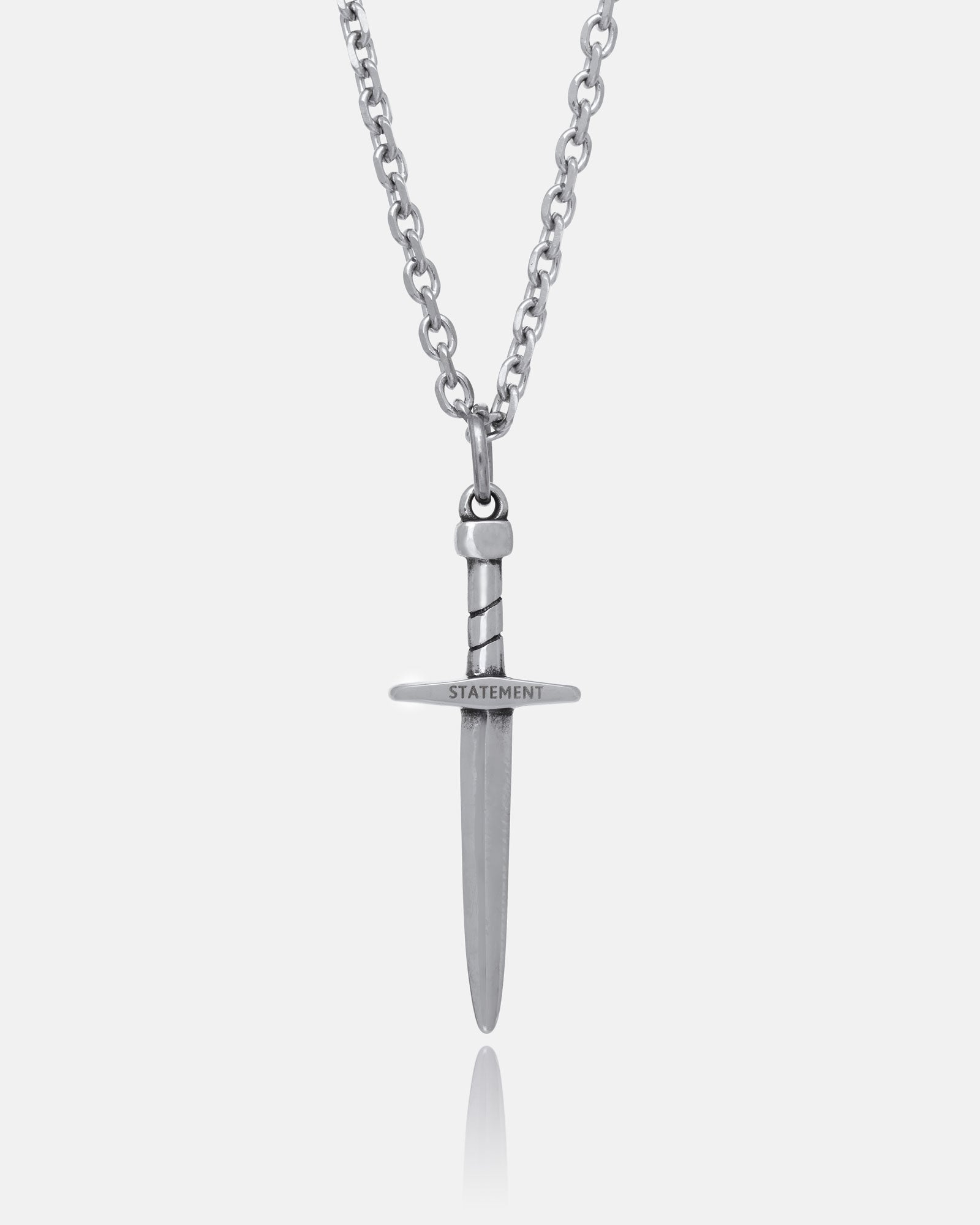 The Dagger Pendant features a hand-carved silver dagger with “STATEMENT” engraved on the crossguard, suspended from a sleek chain and photographed against a plain white background.