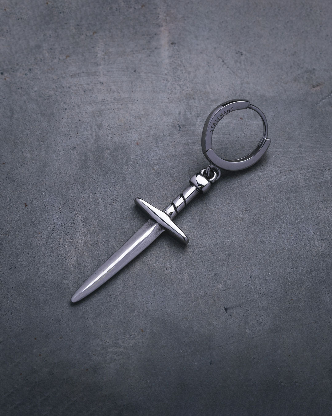 The Dagger Earring—a silver accessory shaped like a small dagger with a straight blade and crossguard, reminiscent of statement jewelry—rests on a textured gray surface.