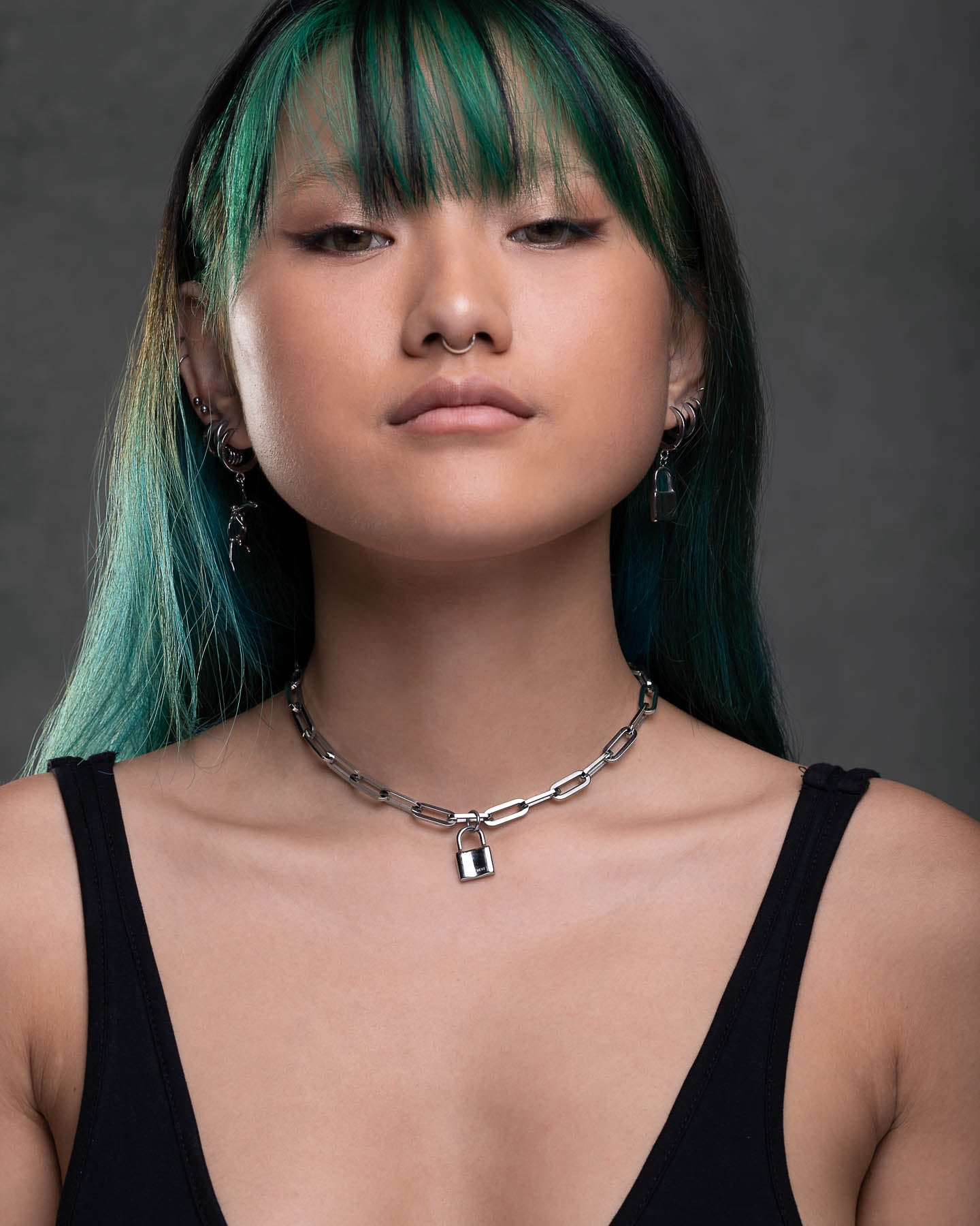 A person with straight teal and black hair and blunt bangs confidently faces the camera against a gray background, wearing silver earrings, a black top, and The Dainty Charm Choker (Custom Pendant Choice).