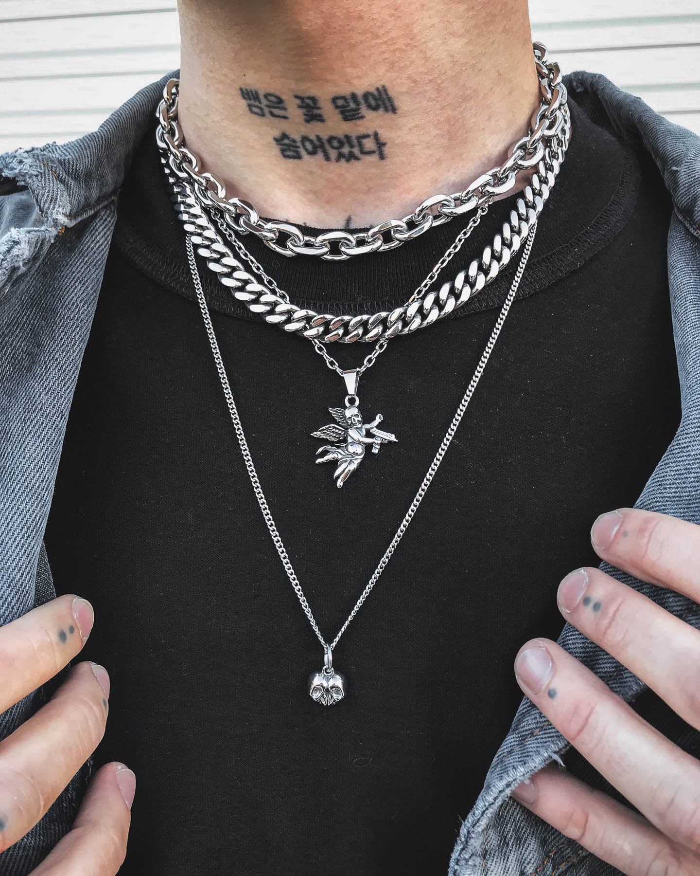 A person wearing the Dead Love Stack layered chain necklace—featuring fairy and skull heart pendants—has a Korean neck tattoo, hand tattoos, and sports a distressed denim jacket over a black shirt.
