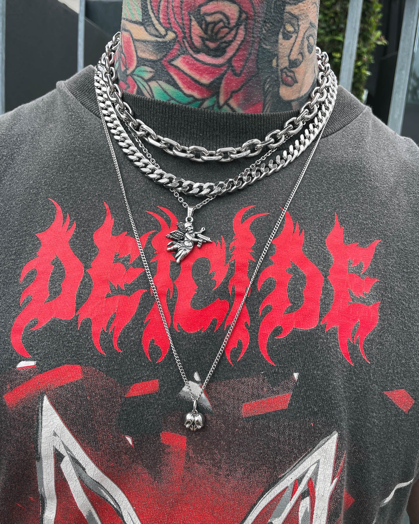 A person wearing the Dead Love Stack grunge necklace set—layered chains over a black shirt with DEICIDE in red, flame-like letters. Colorful tattoos of flowers and a portrait cover their neck and part of their face.