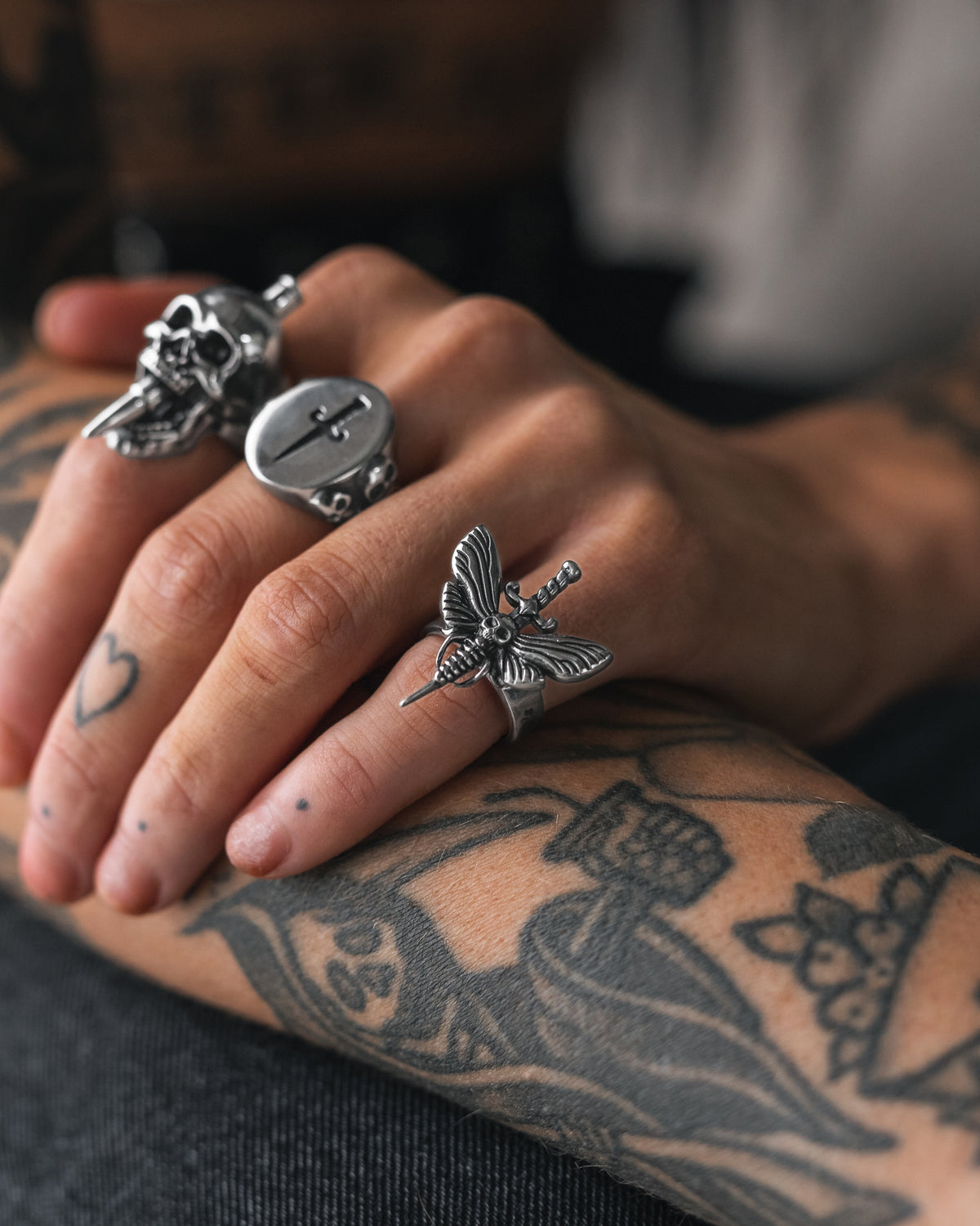 Close-up of a tattooed hand wearing a silver Statement Collective Death’s Head Moth Ring alongside a skull ring and a signet ring with a cross.