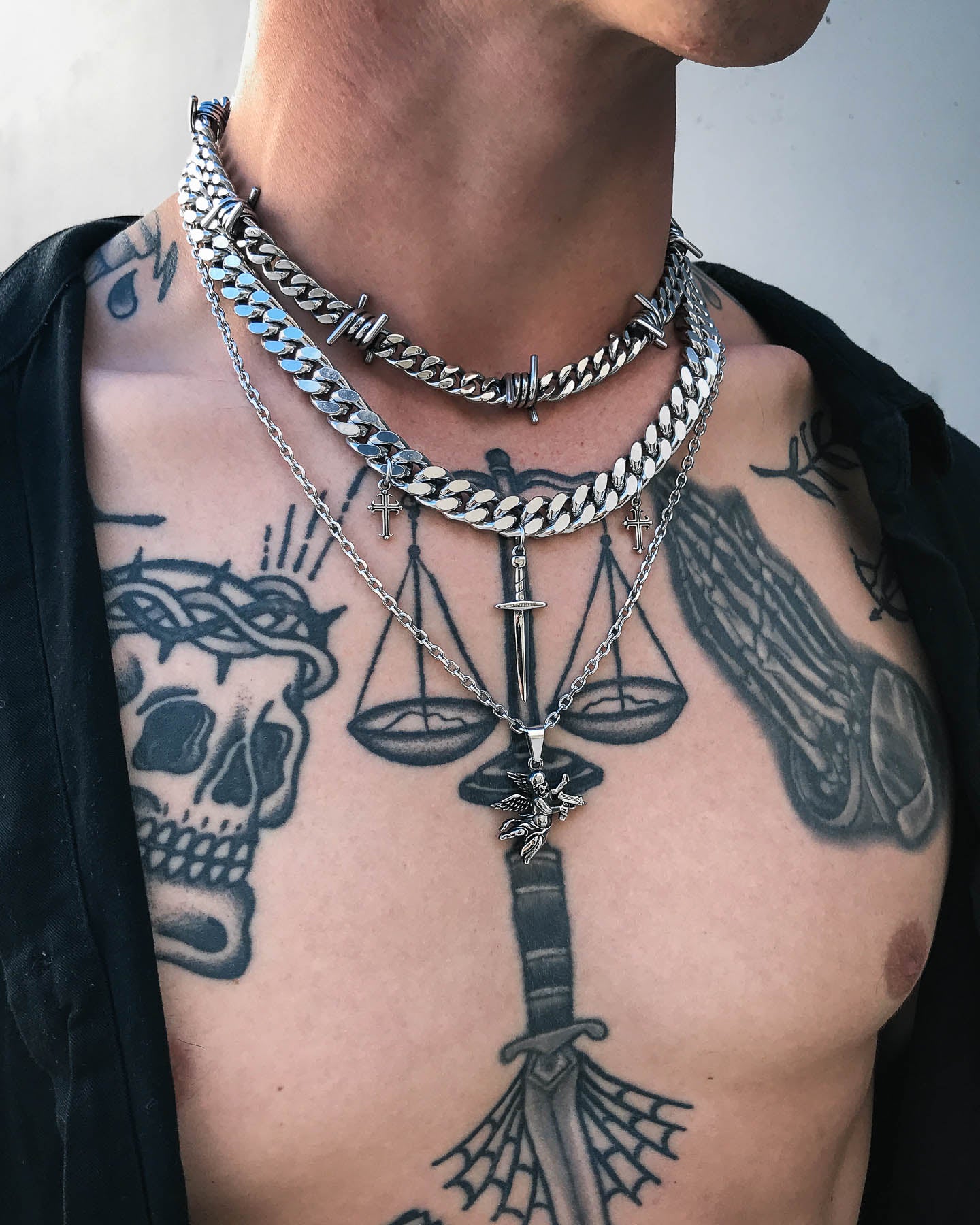 Wearing a black shirt open to display tattoos of a skull, scales of justice, a sword, and webbing, this person layers multiple silver chains and necklaces featuring barbed wire designs and the Death's Cupid Pendant.