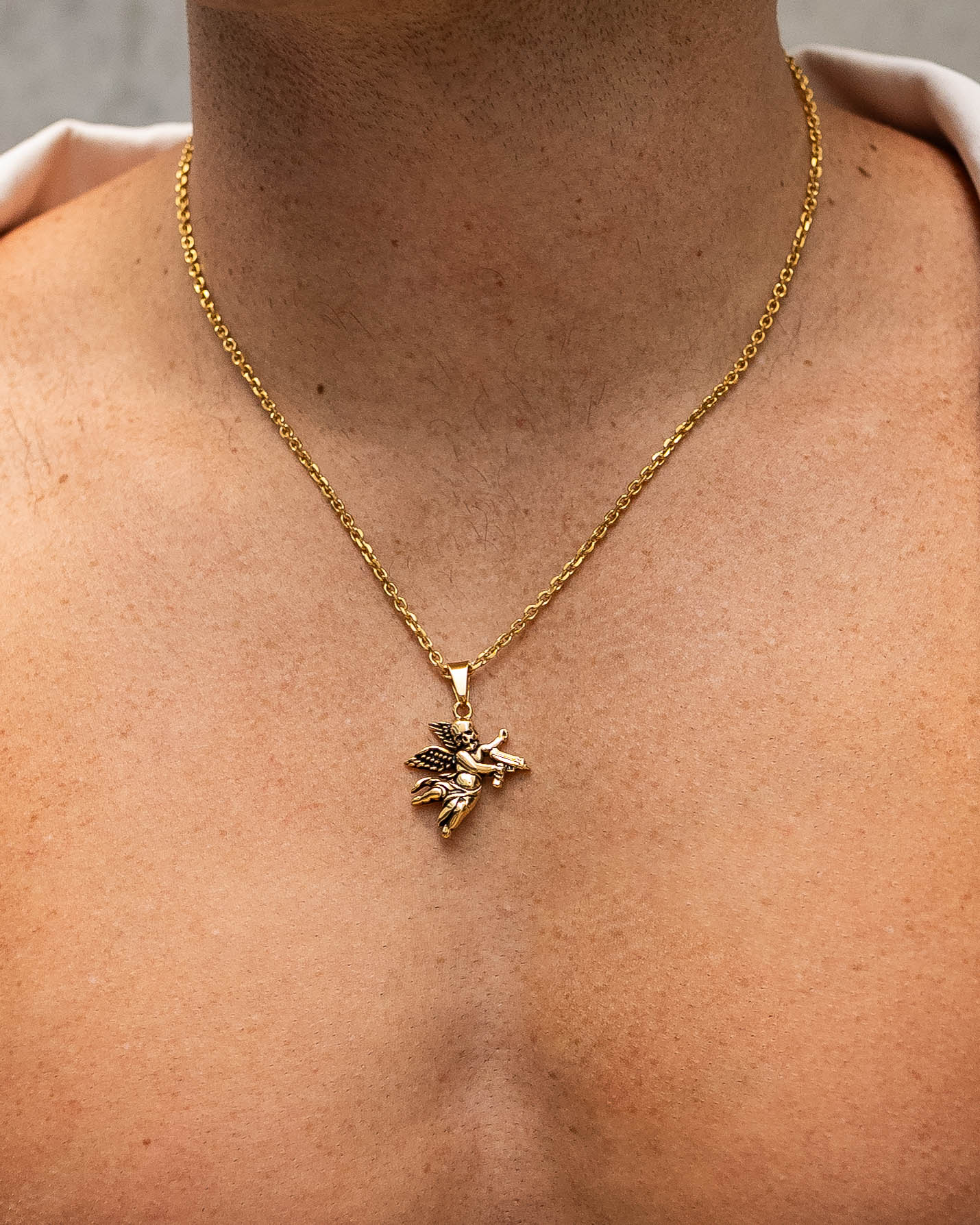 A close-up shows a person's bare chest wearing the Death's Cupid Pendant (Gold), its bee-shaped charm gleaming against light brown skin. The out-of-focus background draws attention to the pendant's elegant shine.