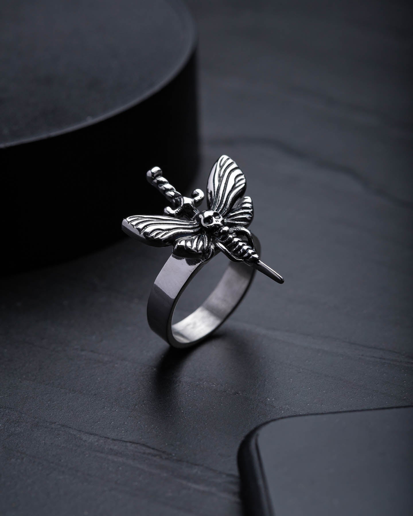 The Death's Head Moth Ring: a silver stainless steel ring featuring a detailed moth and sword design, showcased on a dark textured surface—an exquisite choice for fans of handcrafted jewellery.