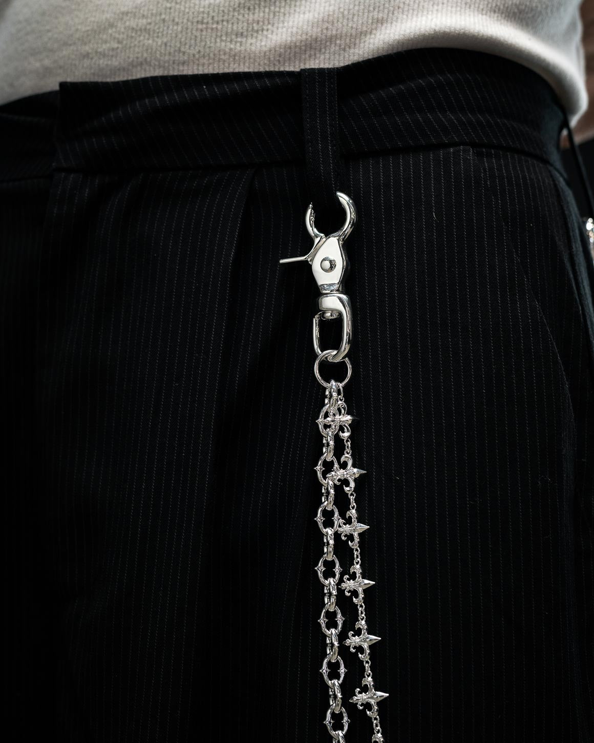 Close-up of black pinstripe pants accessorized with the Statement Collective “Baroque” Fleur De Lis Wallet Chain in sterling silver, clipped to a belt loop and paired with a white textured top.