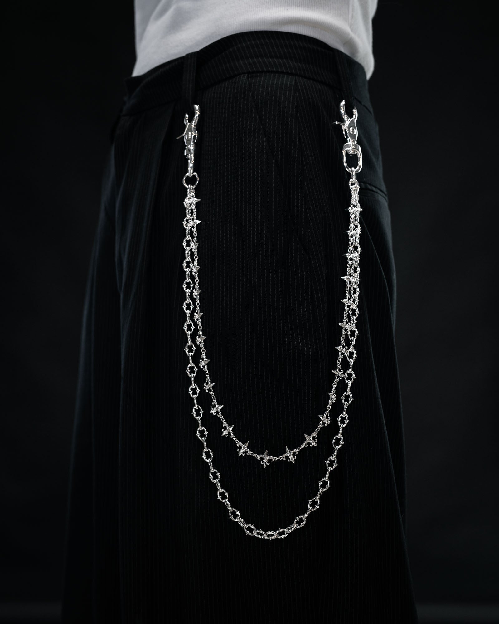 Close-up of a person wearing black pants with the Statement Collective “Baroque” Fleur De Lis Wallet Chain—two sterling silver decorative chains attached to the waistband, hanging in loops on the side like a stylish accessory—set against a dark background.