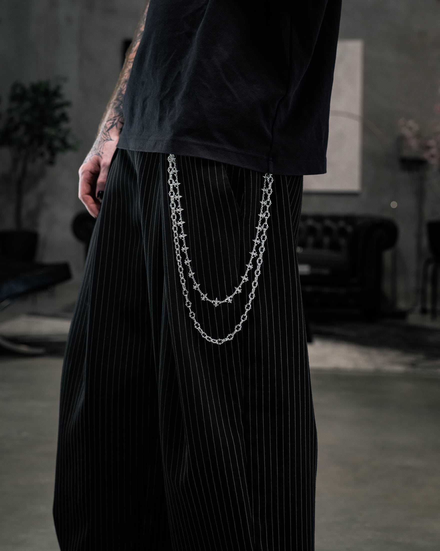 A person wearing black pinstripe pants accessorized with the “Baroque” Fleur De Lis Wallet Chain by Statement Collective, paired with a black t-shirt. The background features a dark sofa and a blurred indoor setting.