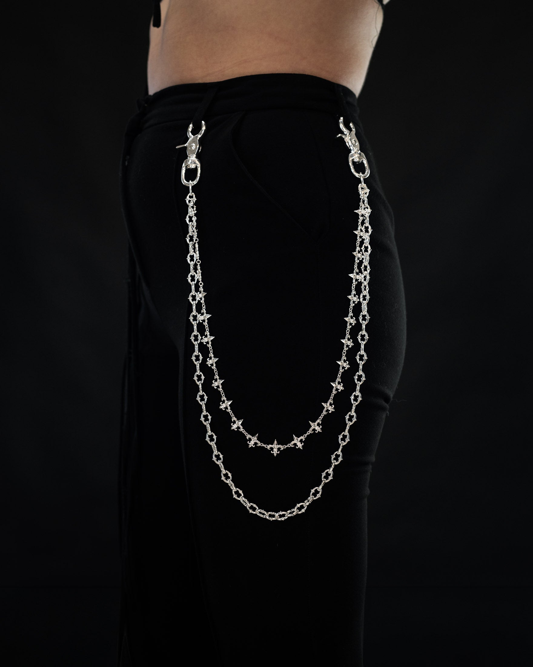 A person wearing black pants accessorized with the Statement Collective “Baroque” Fleur De Lis Wallet Chain, featuring a double sterling silver design clipped to the waistband, against a black background. Only the lower torso and upper thigh are visible.
