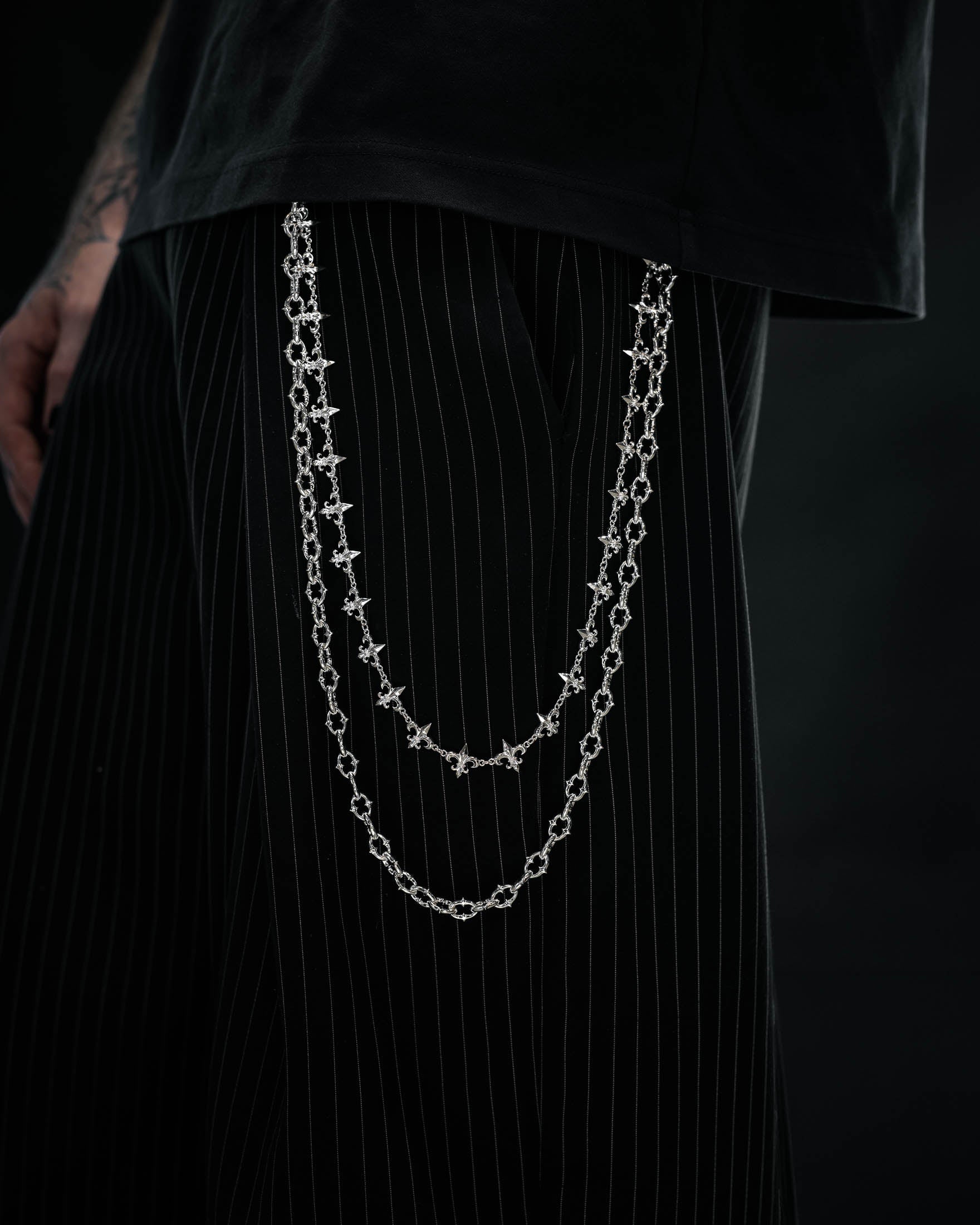A person wearing black pinstripe pants and a black shirt showcases the Statement Collective “Baroque” Fleur De Lis Wallet Chain, its intricate sterling silver links draped from their pocket. Only the lower torso and part of one hand are visible.
