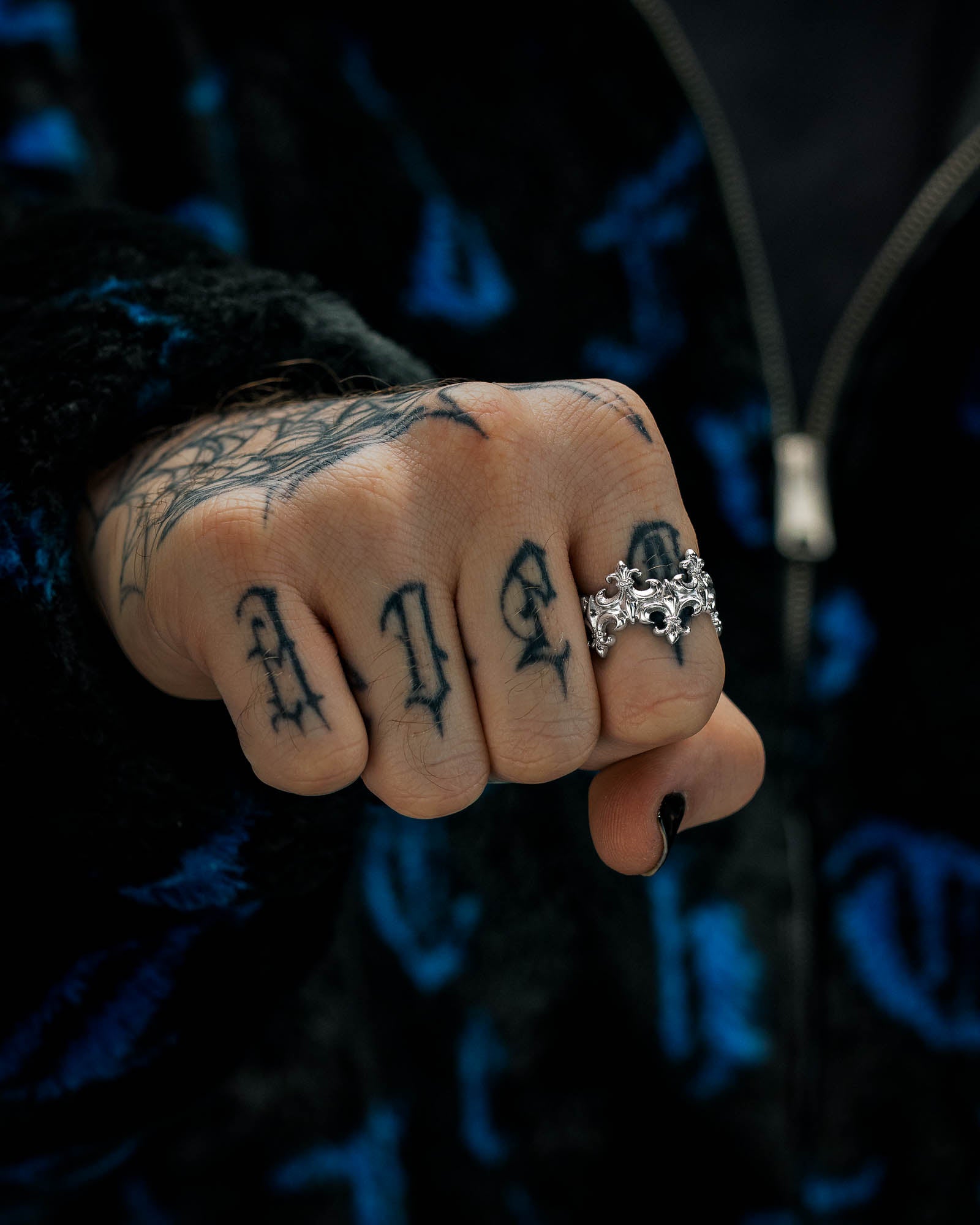 A close-up of a clenched fist with tattoos on the fingers, showcasing the “Crested” Fleur De Lis Ring by Statement Collective—a detailed 316L stainless steel band styled like a crown—alongside a spiderweb tattoo on the hand. The person is dressed in a dark, patterned jacket.
