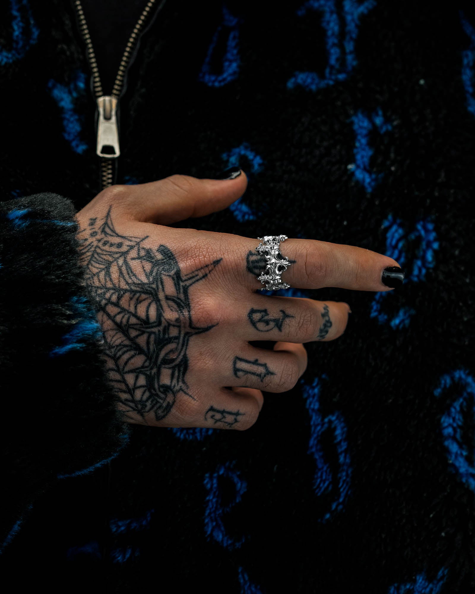 A tattooed hand with black nails points, adorned with the Statement Collective “Crested” Fleur De Lis ring alongside a silver skull ring. The person is wearing a dark, fuzzy jacket featuring a blue pattern and a visible zipper.