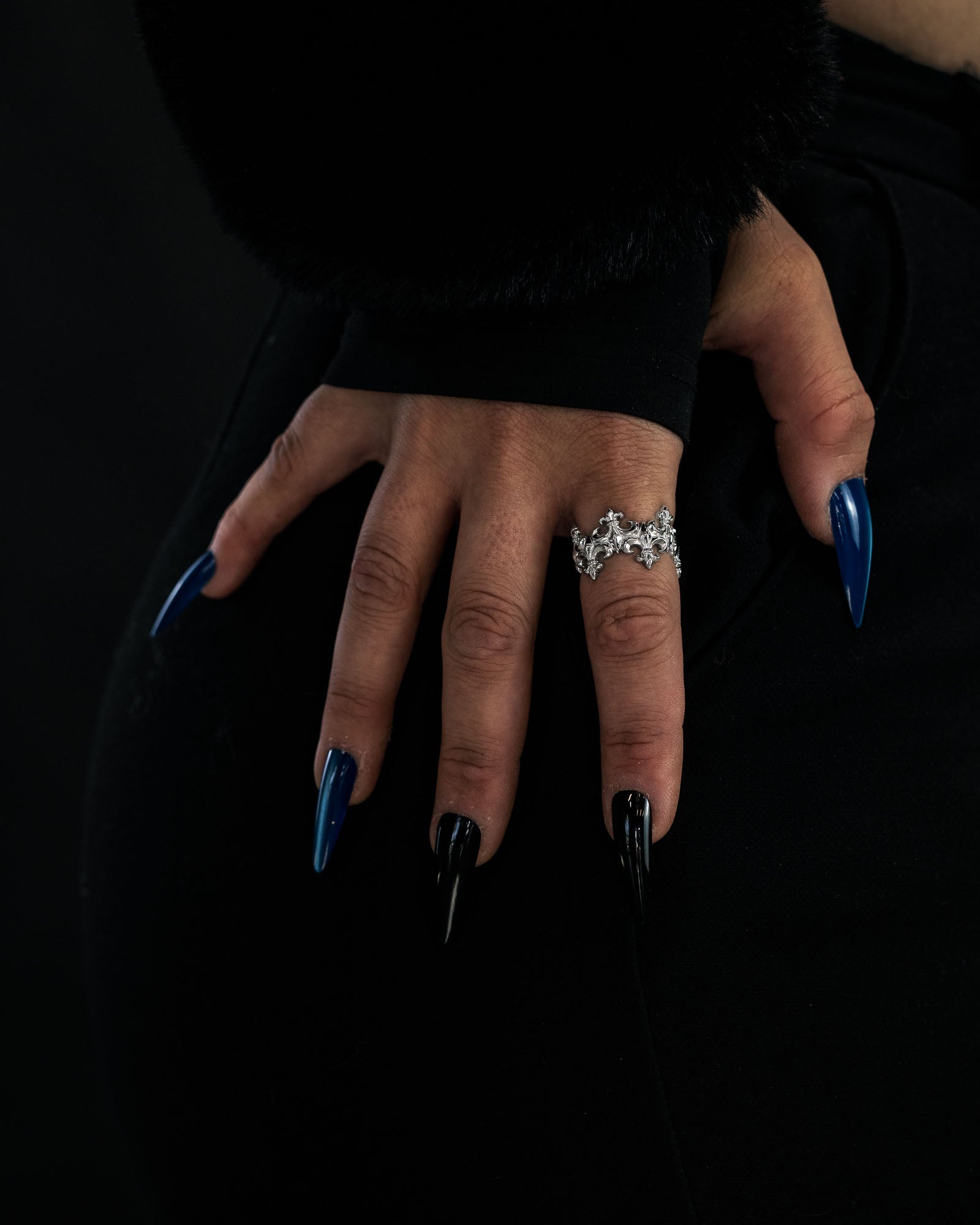 A hand adorned with long, pointed black and blue nails rests on a hip. The person wears a chunky silver ring shaped like leaves, paired with Statement Collective’s striking “Crested” Fleur De Lis Ring, which features a distinctive crown-like band. Black clothing and background create a dramatic contrast.