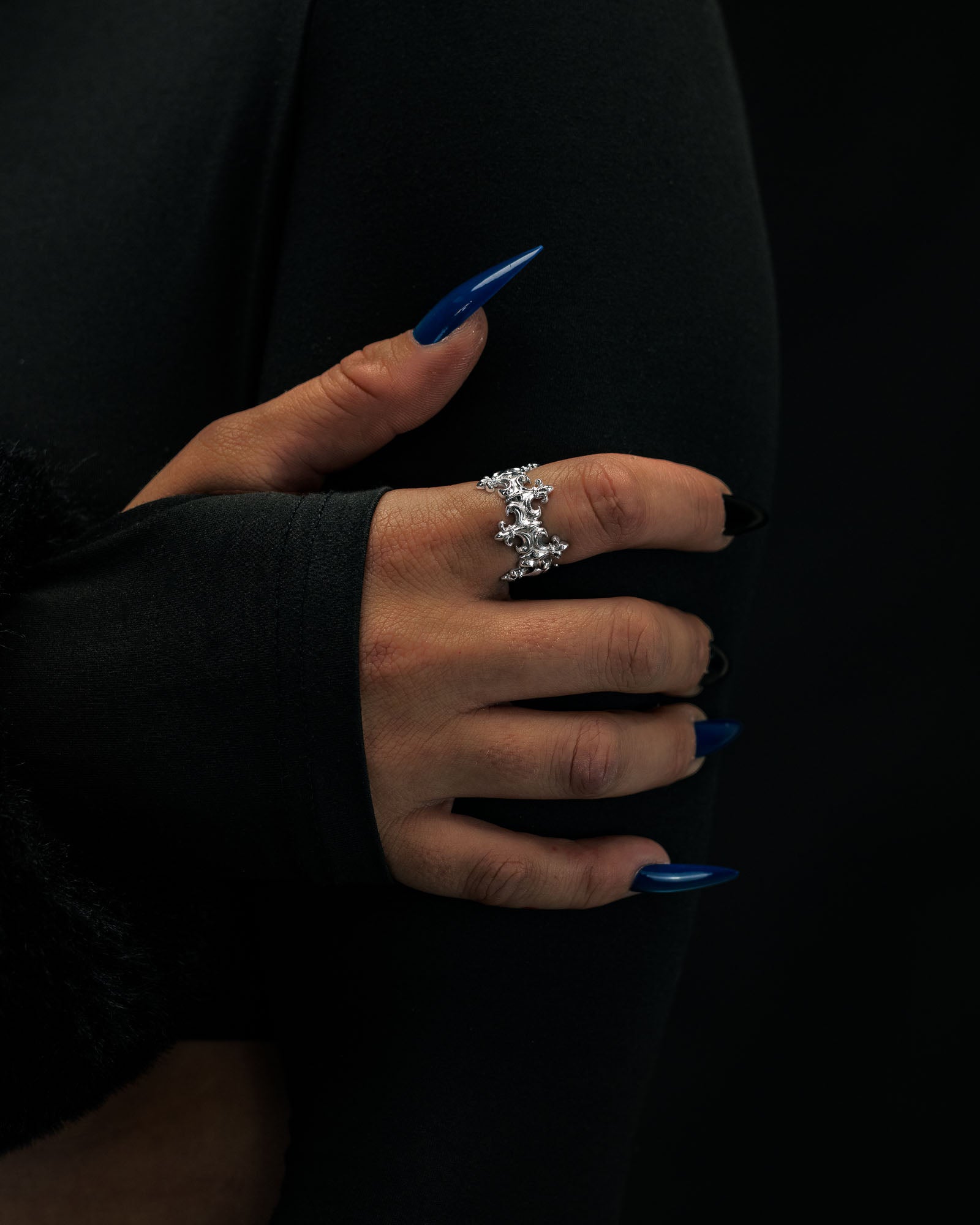 A hand with long, blue-painted nails wears the sparkling “Crested” Fleur De Lis Ring by Statement Collective—a crown-like band. The person, dressed in black with a thumbhole sleeve partially covering their hand, is set against a dark background.