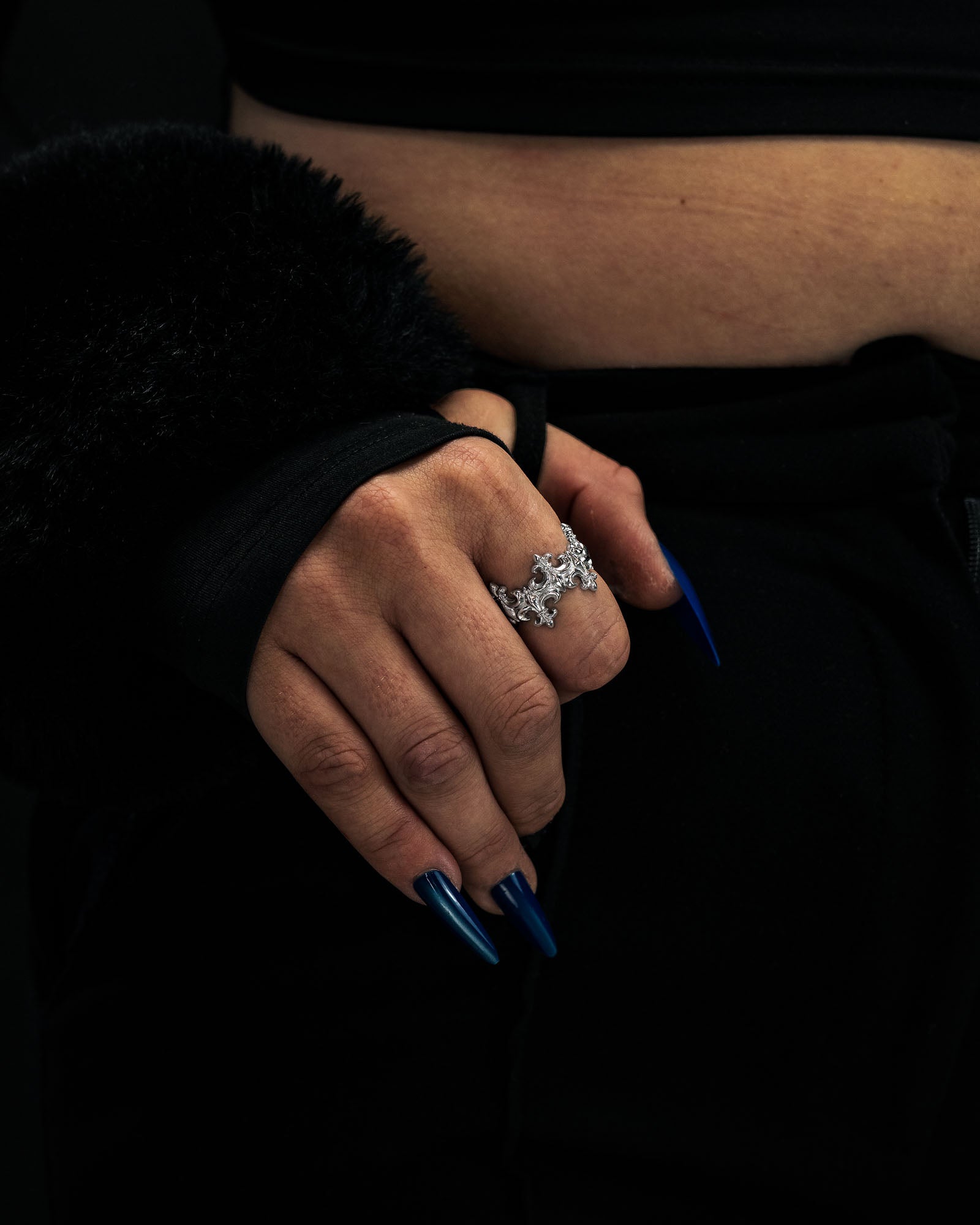 A hand with long, pointed blue nails wears the “Crested” Fleur De Lis Ring by Statement Collective, crafted from 316L stainless steel. The person is dressed in black, with a portion of their midriff and a fuzzy sleeve visible against a dark background; the crown-like band of the ring lends an extra touch of elegance to this striking look.