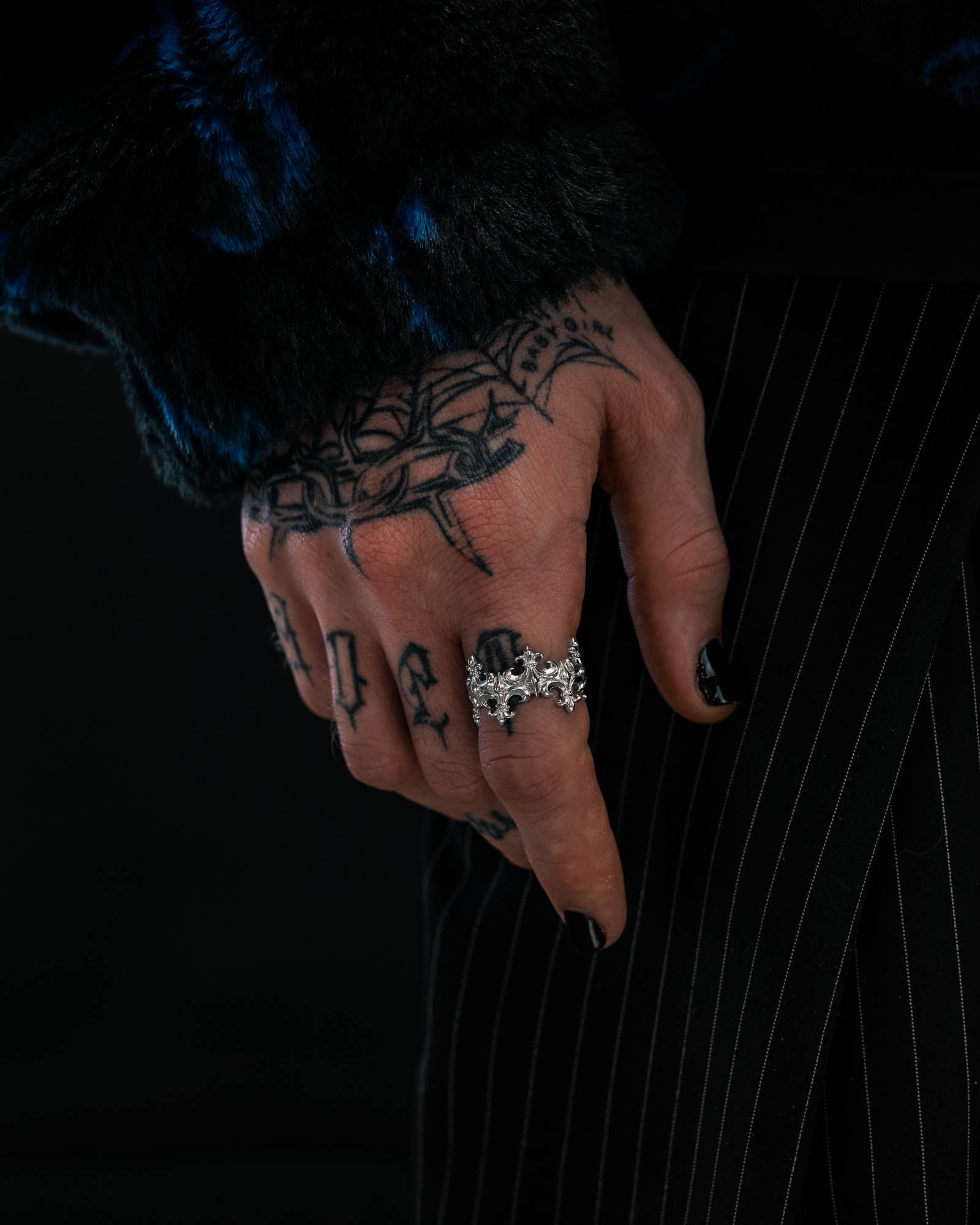 A tattooed hand with black-painted nails displays a detailed silver bat-shaped ring alongside the eye-catching “Crested” Fleur De Lis Ring by Statement Collective. The wearer’s dark fuzzy sleeve and pinstriped pants stand out against a black background, enhancing the bold aesthetic.