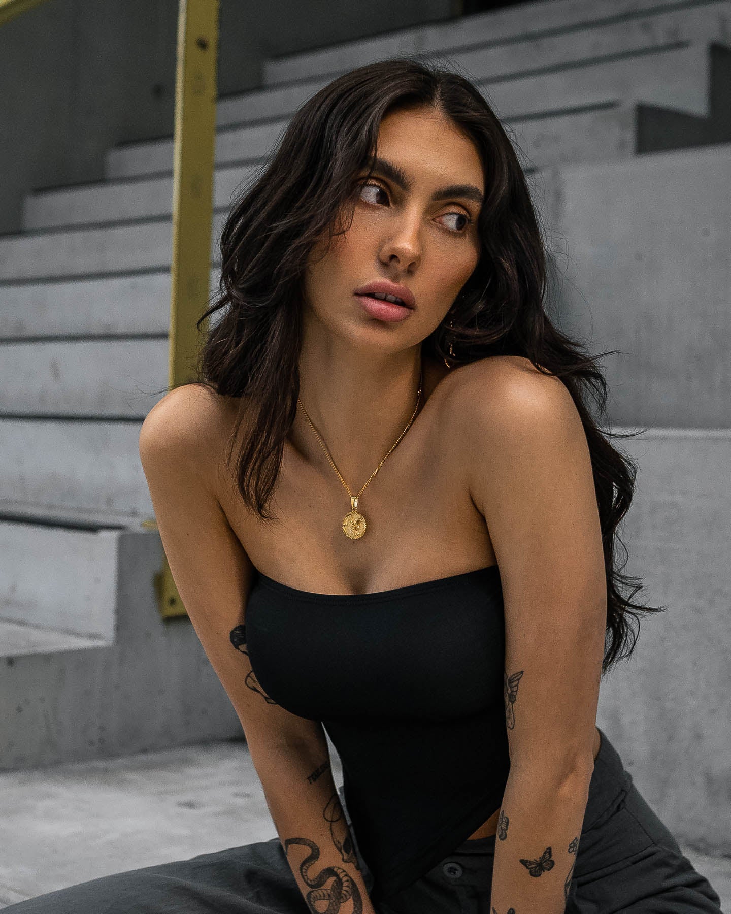 A woman with long dark hair, in a black strapless top and the Fallen Angel Medallion Pendant (Gold), sits on concrete steps, gazing thoughtfully to her left. Tattoos are visible on her arms.