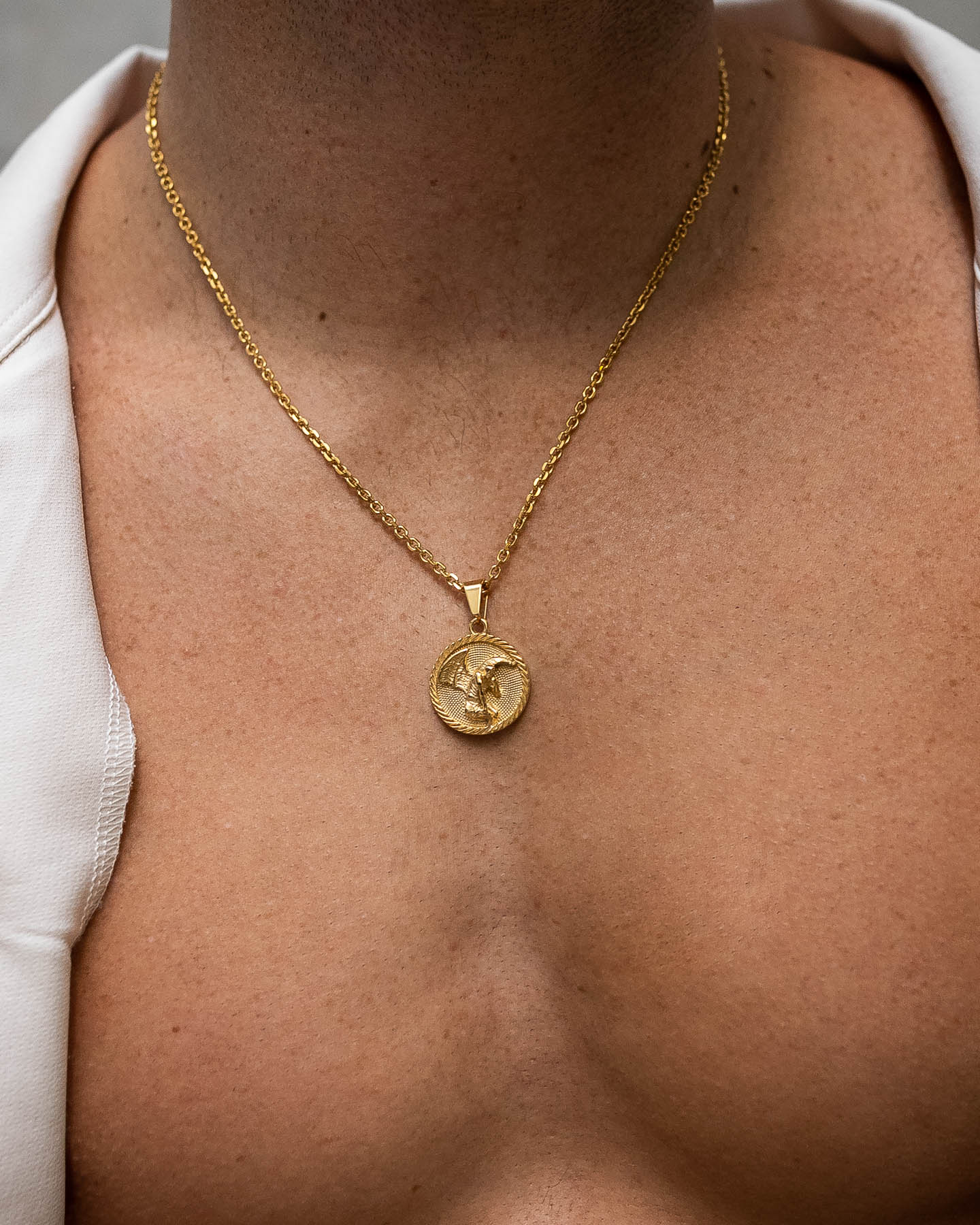 A close-up of a bare chest wearing the Fallen Angel Medallion Pendant (Gold), an 18k gold, non-tarnish, waterproof necklace with a raised winged figure, shining beneath a partially open white shirt.