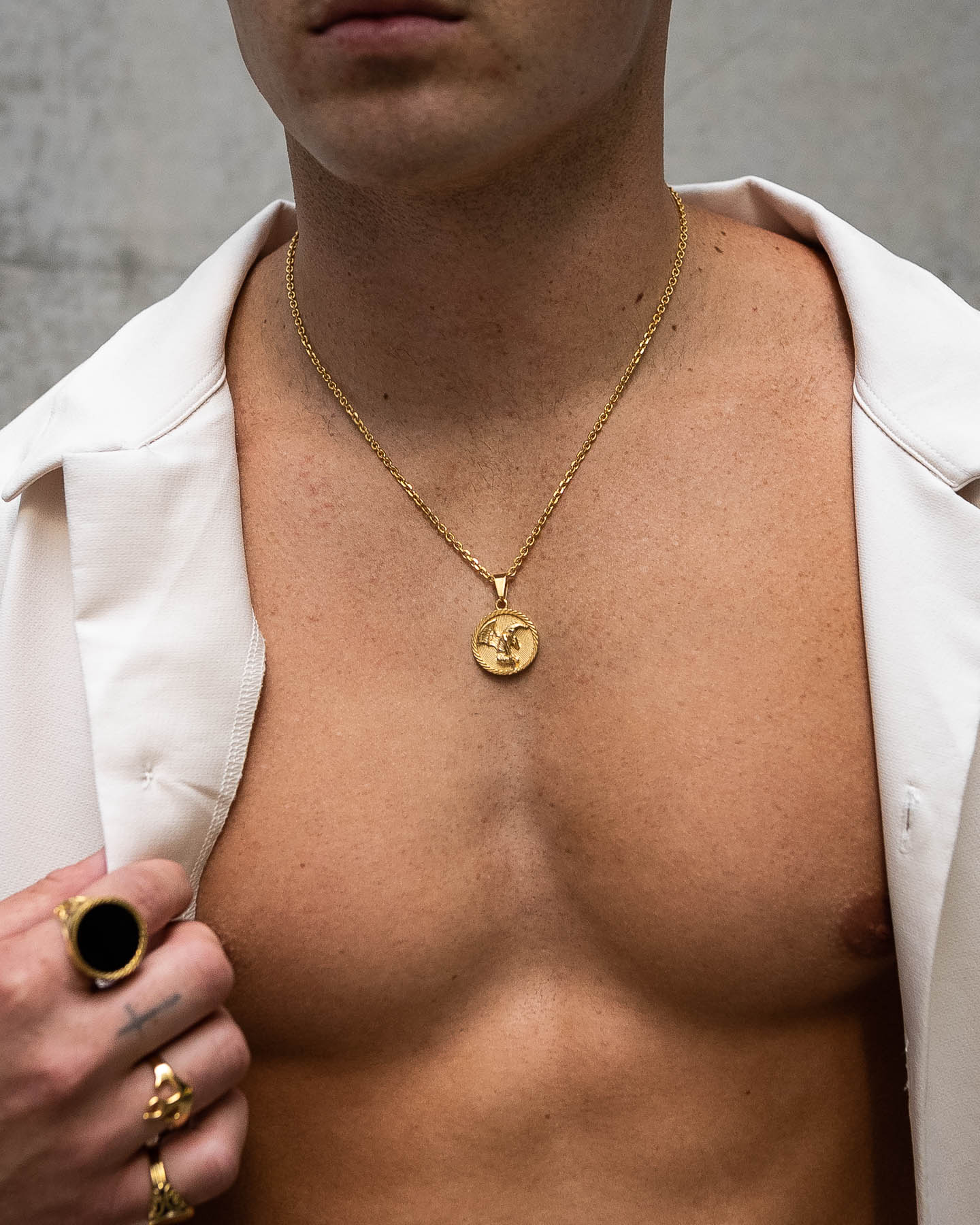 A shirtless man in an open white shirt showcases the Fallen Angel Medallion Pendant (Gold) and several waterproof, non-tarnish rings—including one with a black stone—against a neutral, blurred background.