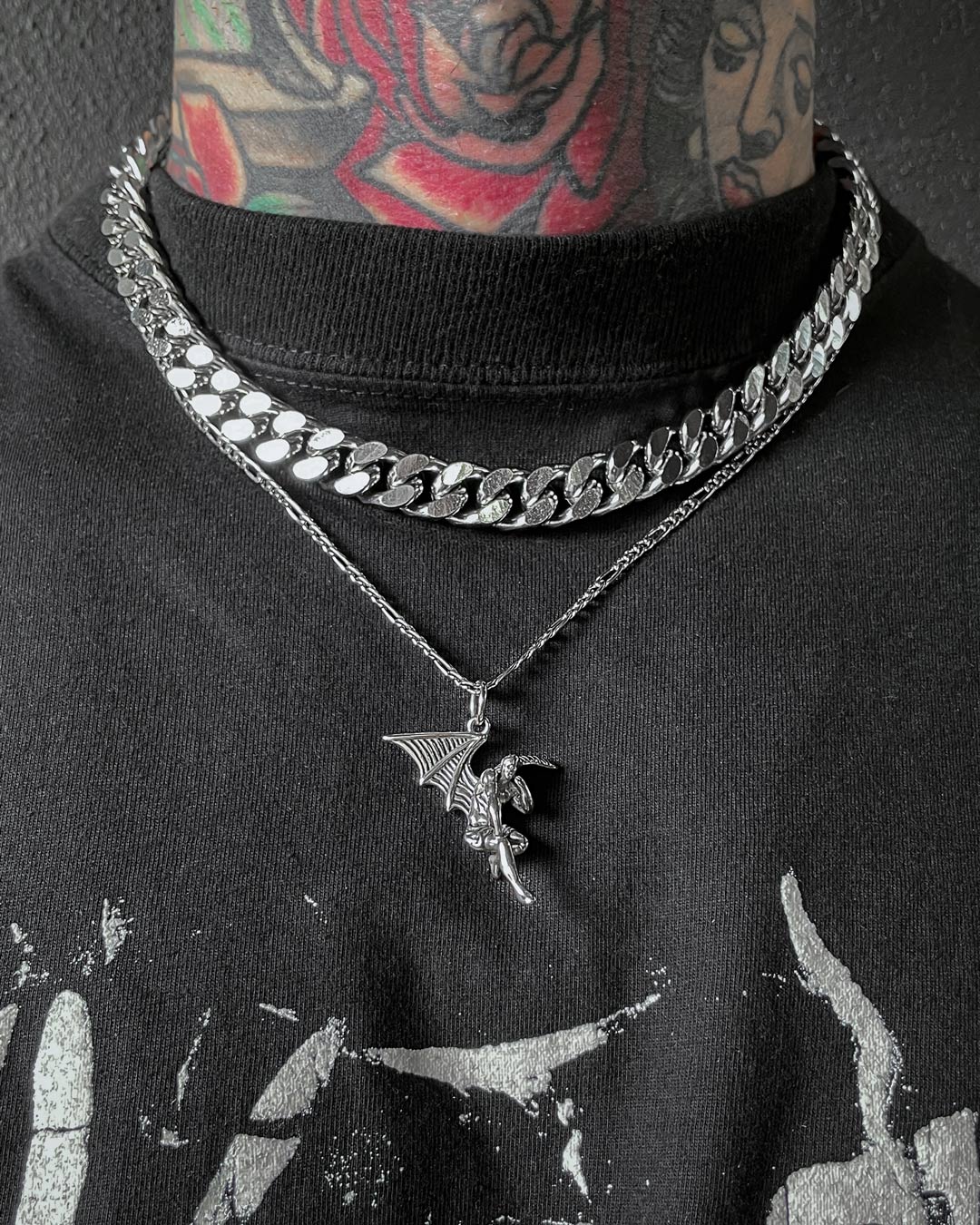 A person with colorful neck tattoos wears two silver necklaces—a thick Cuban link chain and another with the Fallen Angel Pendant—paired with a black graphic t-shirt that highlights bold jewellery designs.