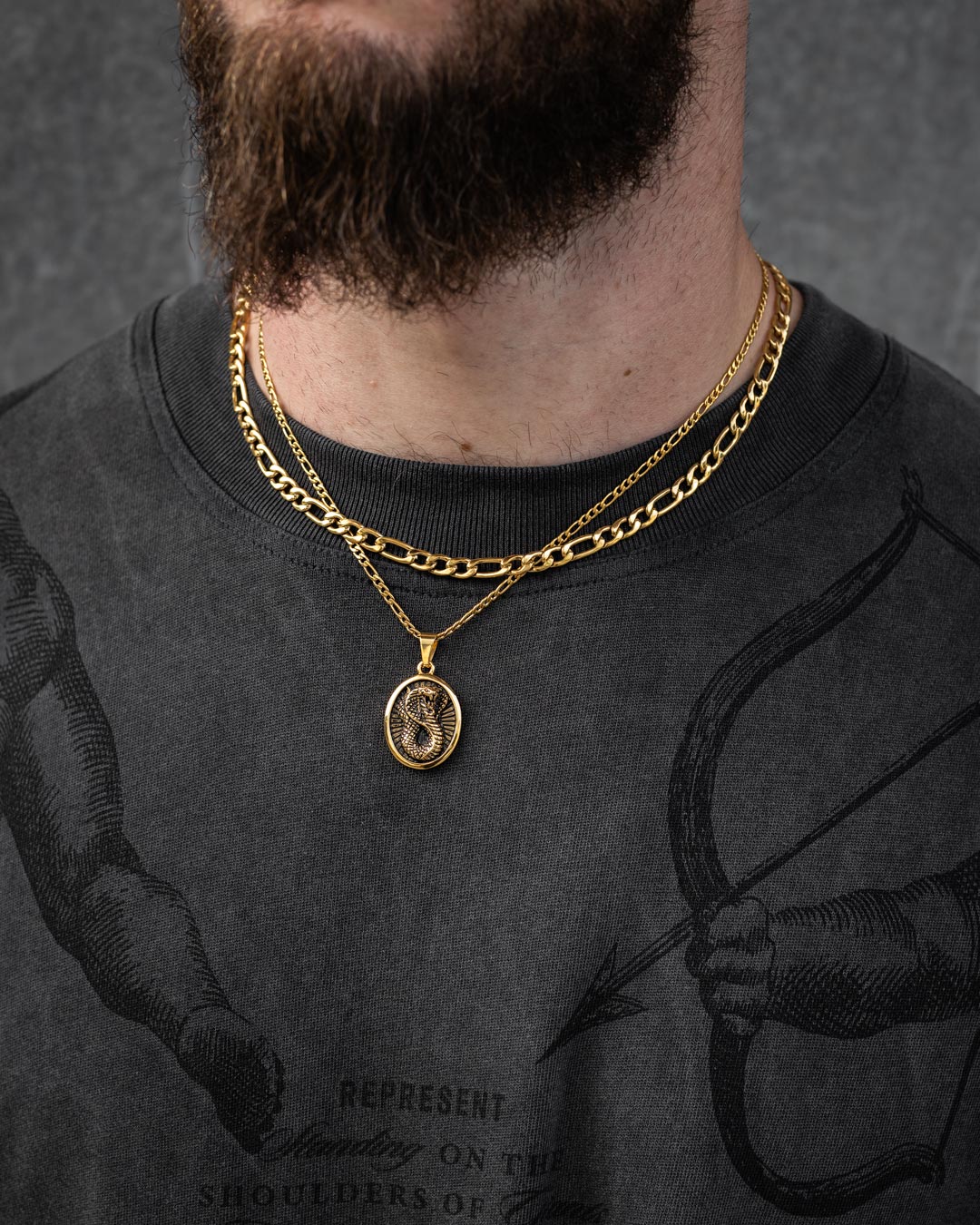 A close-up of a bearded man wearing Statement Collective’s Figaro Set (Custom Pendant Choice) (Gold) over a black graphic T-shirt with a drawing of an arm holding a bow and an inscription.