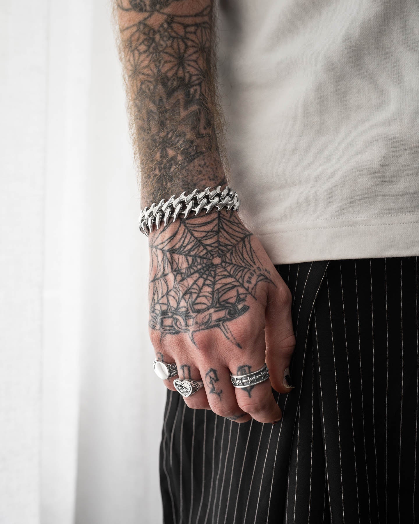 A tattooed hand adorned with silver rings and the bold "Fishbone" Spiked Cuban Bracelet in 316L stainless steel rests by their side. Dressed in a white shirt and black pinstriped pants, they showcase a prominent spiderweb tattoo design.