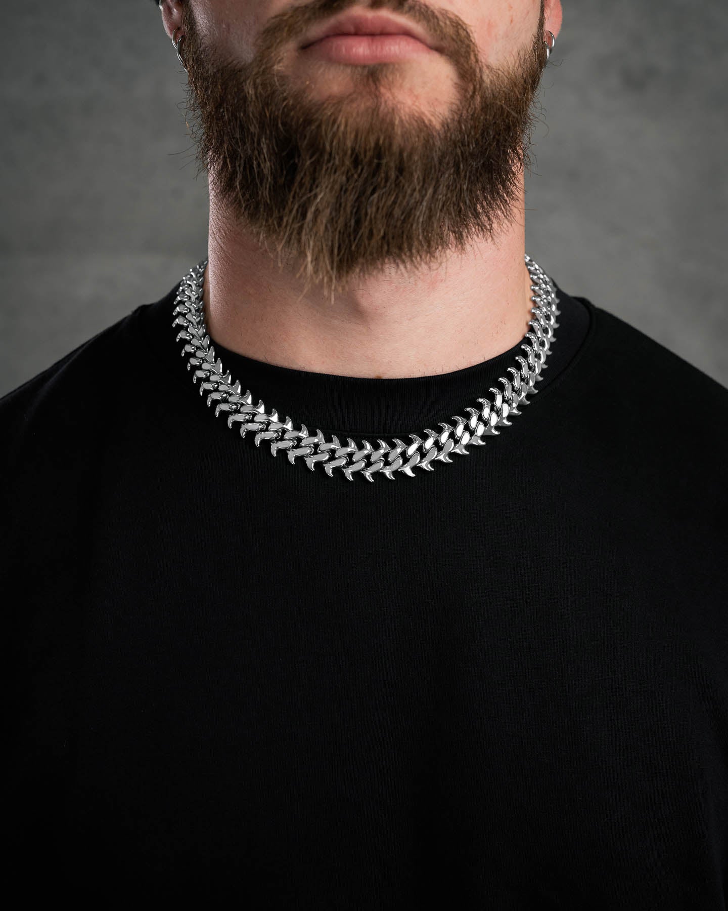 A man with a full brown beard in a plain black shirt wears the "Fishbone" Curved Spike Cuban Chain, photographed against a gray background with his face partially out of frame.