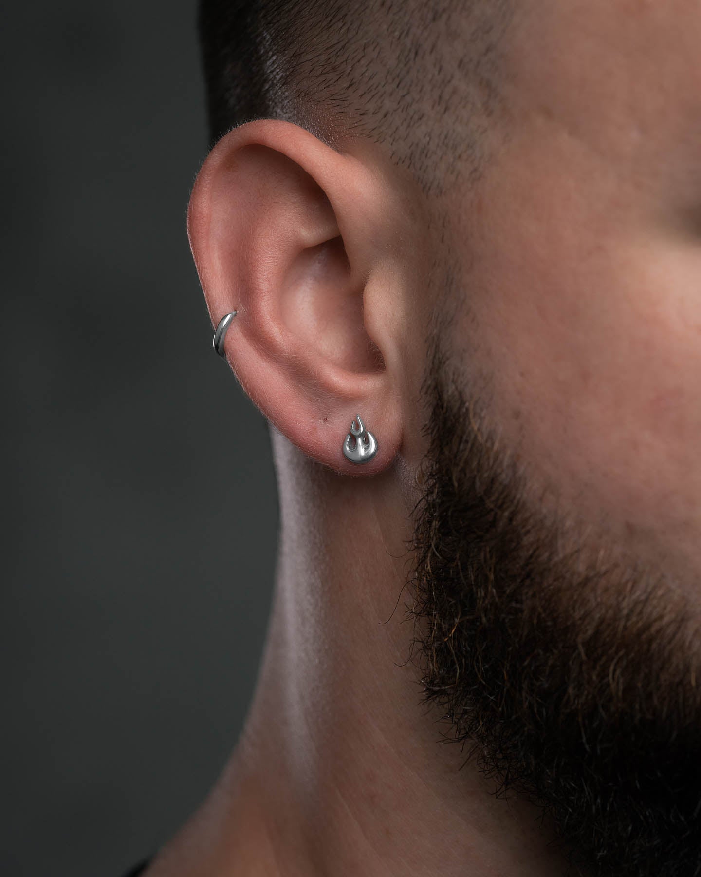Close-up of a person with short hair and a beard wearing the Flame Stud Earring in the earlobe. The stainless steel design stands out against a dark, blurred background.