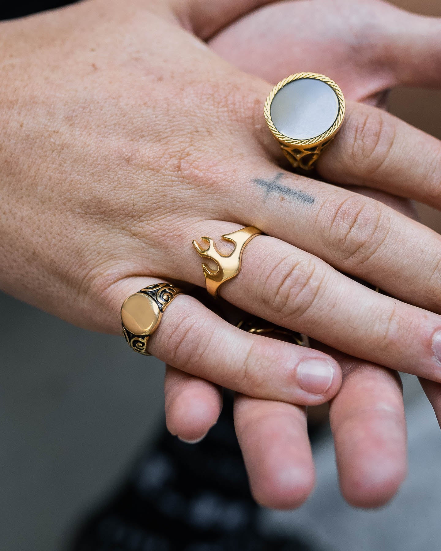 Two hands display several gold pieces, featuring the Flame Ring (Gold), an 18k signet ring, and an engraved band. A small cross tattoo appears on one finger.