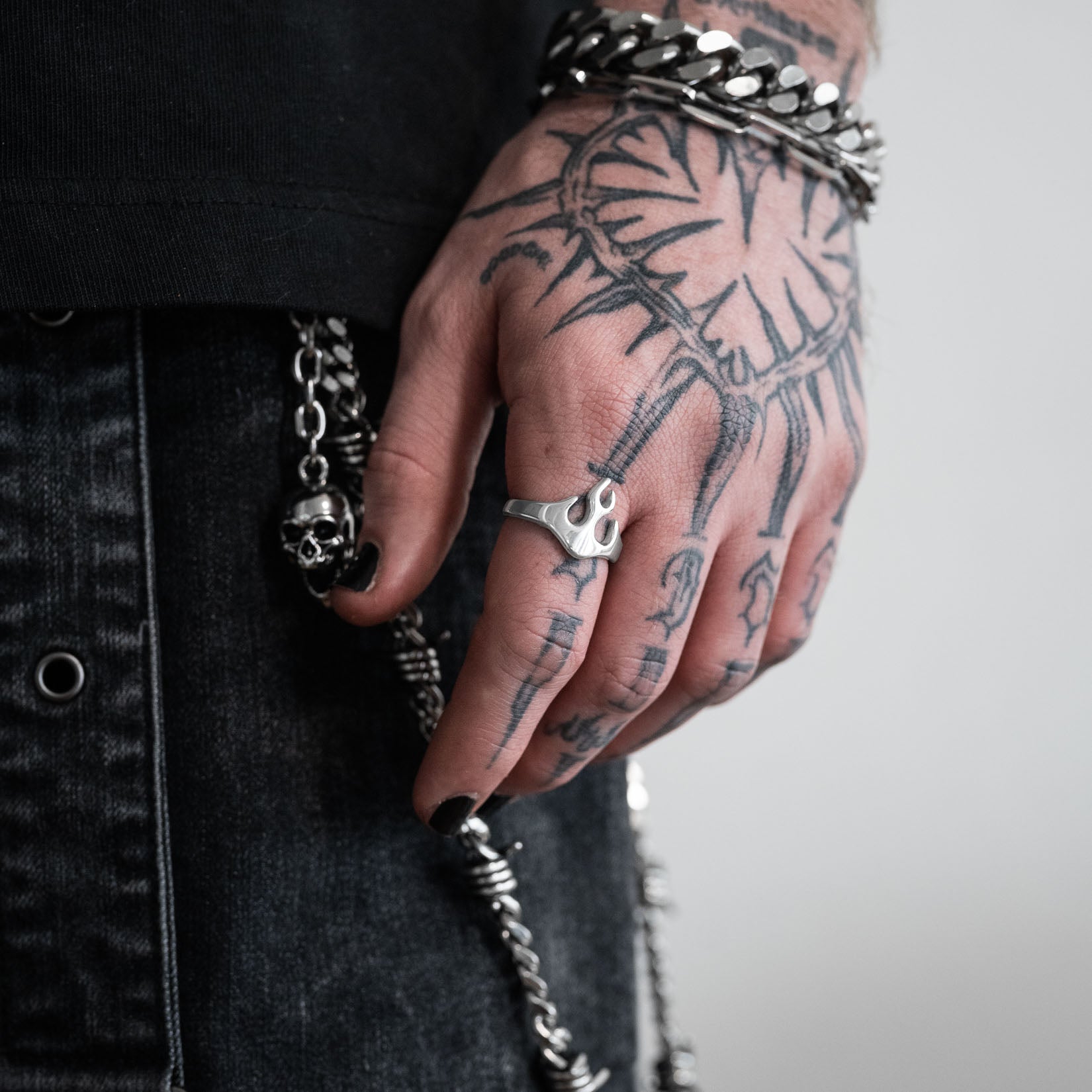 Hand with tattoos wearing a silver flame shaped ring and black nail polish, with a blurred background.