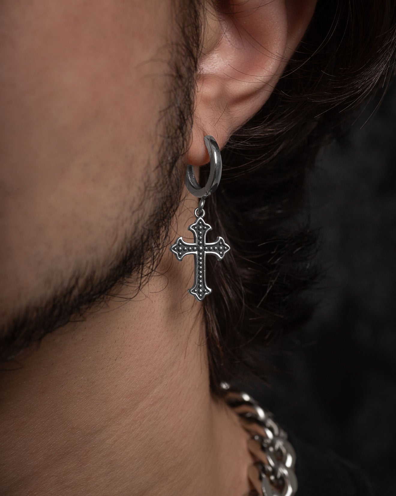 A close-up of a person’s ear wearing the French Cross Earring—a metallic hoop with a dangling cross pendant. The photo also shows part of their beard, hair, and a chunky chain necklace.