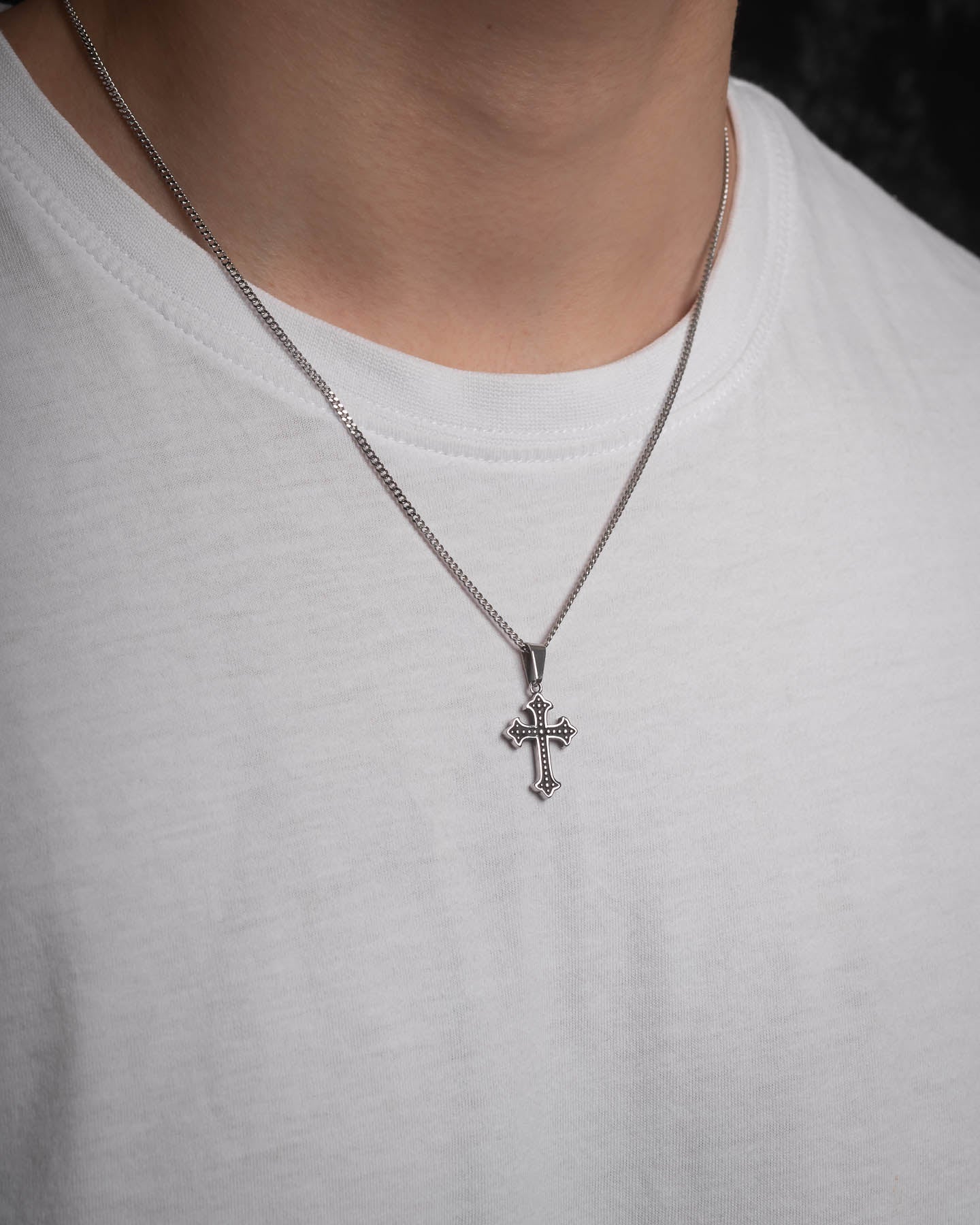 A person wears a white t-shirt and the French Cross Pendant, a silver figaro chain necklace with a detailed cross, with focus on the neck and upper chest area.