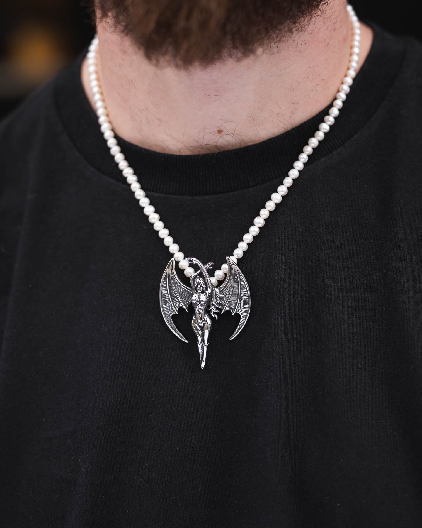 Someone wears the Freshwater Pearl Necklace With Custom Pendant Choice, featuring a silver winged humanoid pendant, over a plain black shirt.