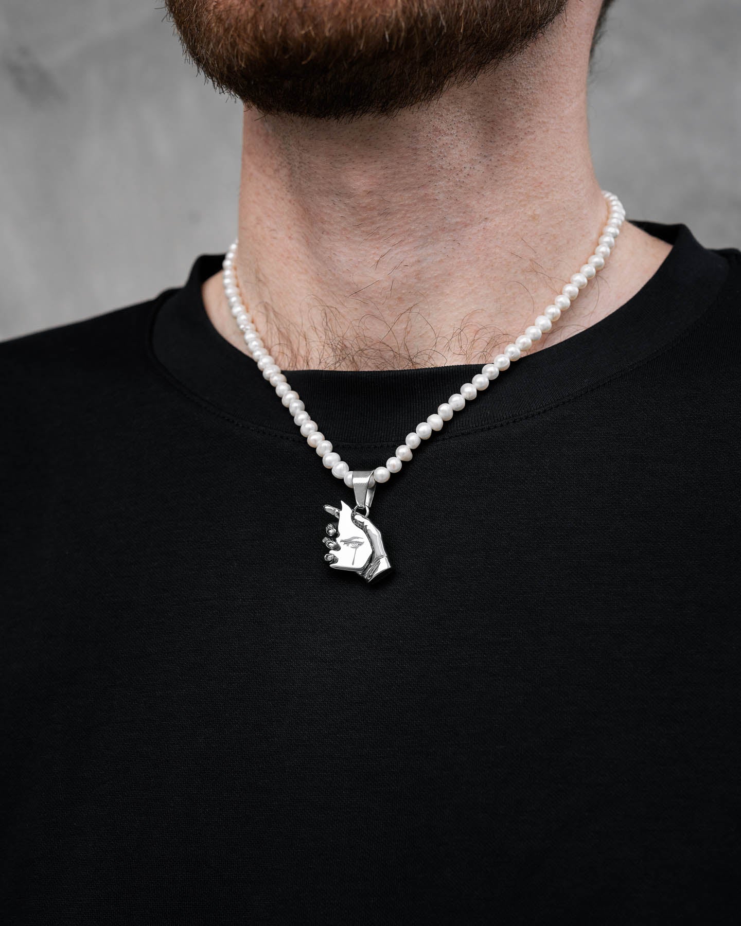 A person wears a black shirt and the Freshwater Pearl Necklace With Custom Pendant Choice, featuring a beaded faux pearl chain and a silver hand peace sign pendant, shown against a neutral gray background.