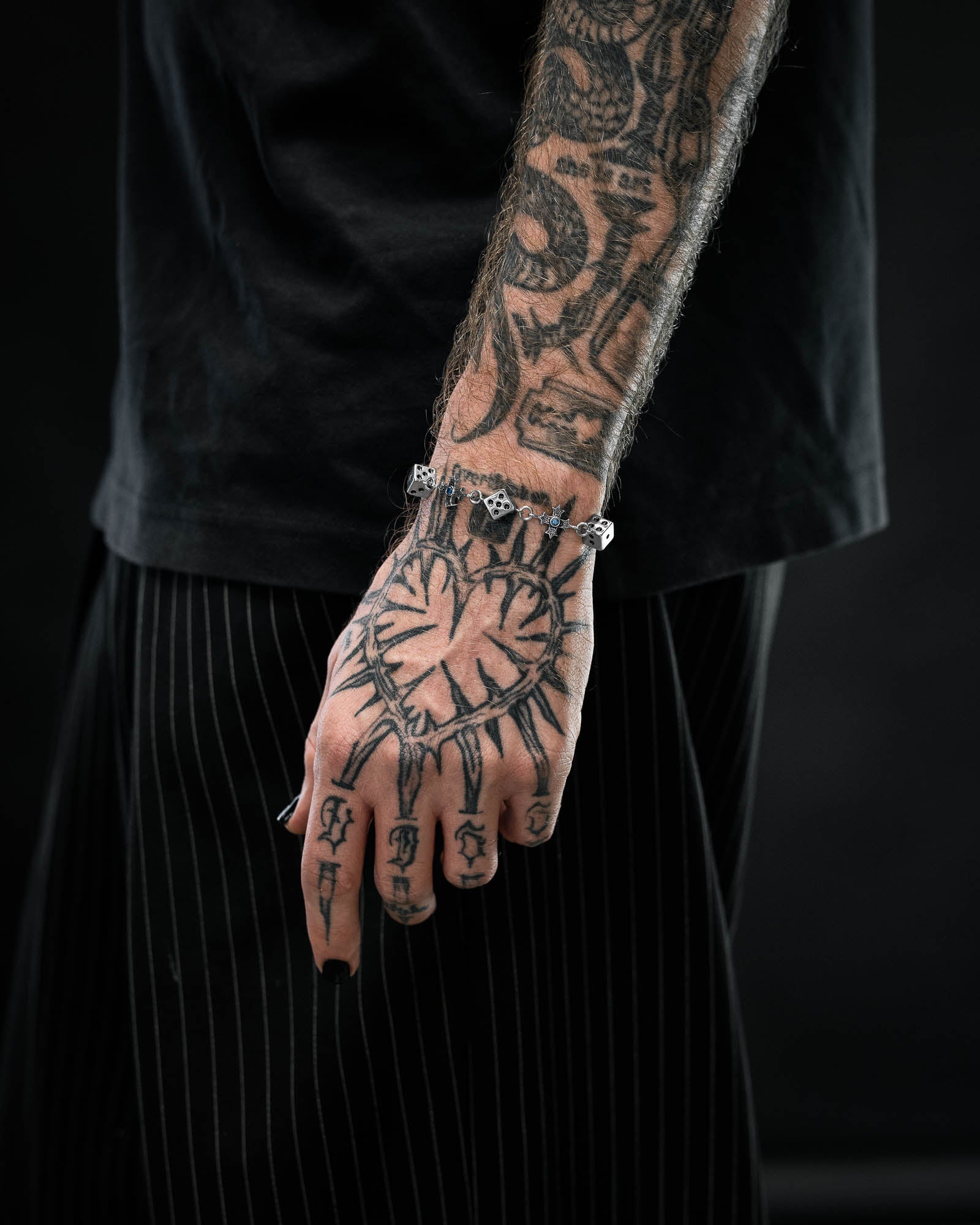 A tattooed arm and hand with a sunburst design showcase the Gambler's Faith Bracelet by Statement Collective. The person wears black pinstripe pants and a black shirt, set against a dark background.