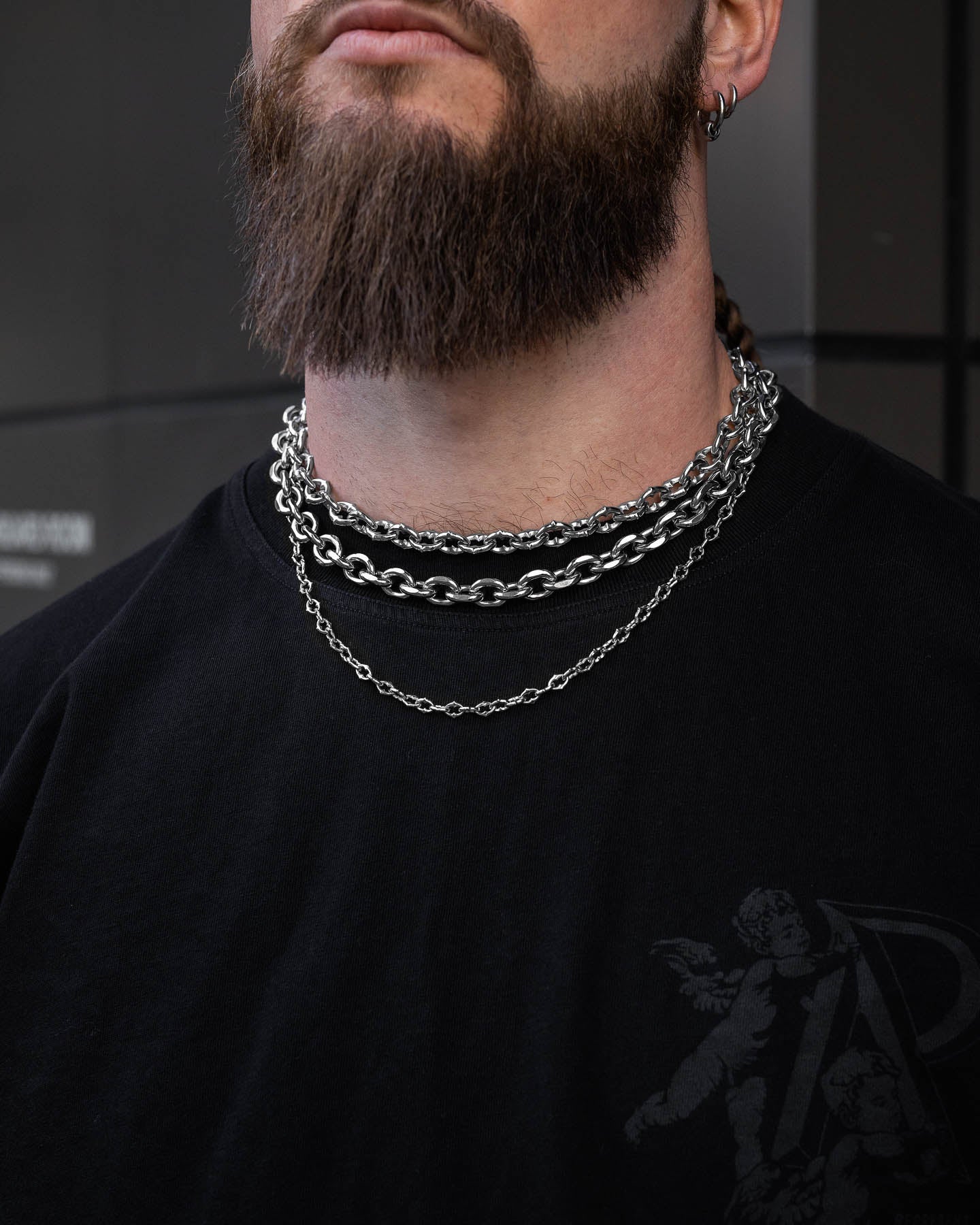 A person with a full beard wears the Go To Church Stack—layered silver chains with a bold cable chain—around their neck and a black t-shirt featuring a small cherub design. The background shows dark, blurred panels.