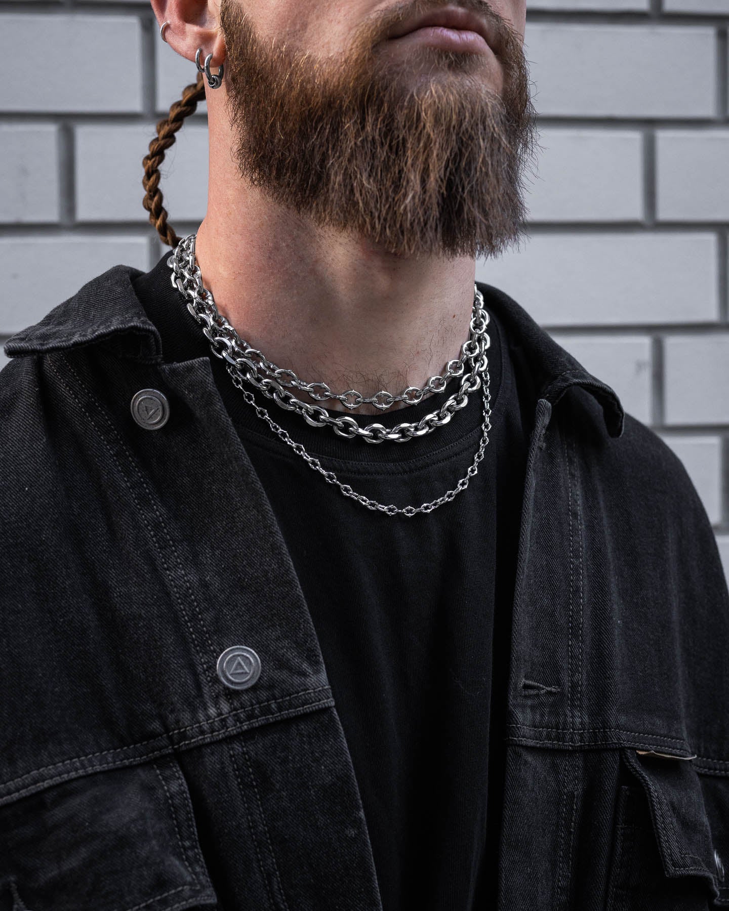 A person with a long beard wears the Go To Church Stack, a striking set of layered silver chains, along with a black denim jacket and shirt, standing in front of a white brick wall.