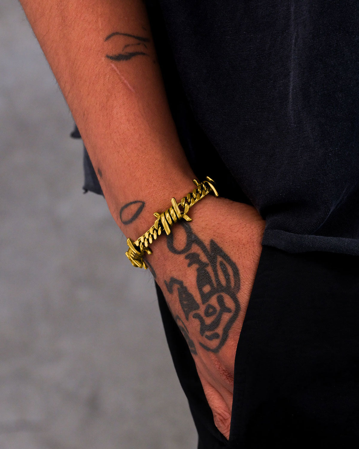 A tattooed individual wears the 6mm Barbed Wire Cuban Bracelet (Gold), their hand in a black pants pocket, making a bold style statement against a neutral background.
