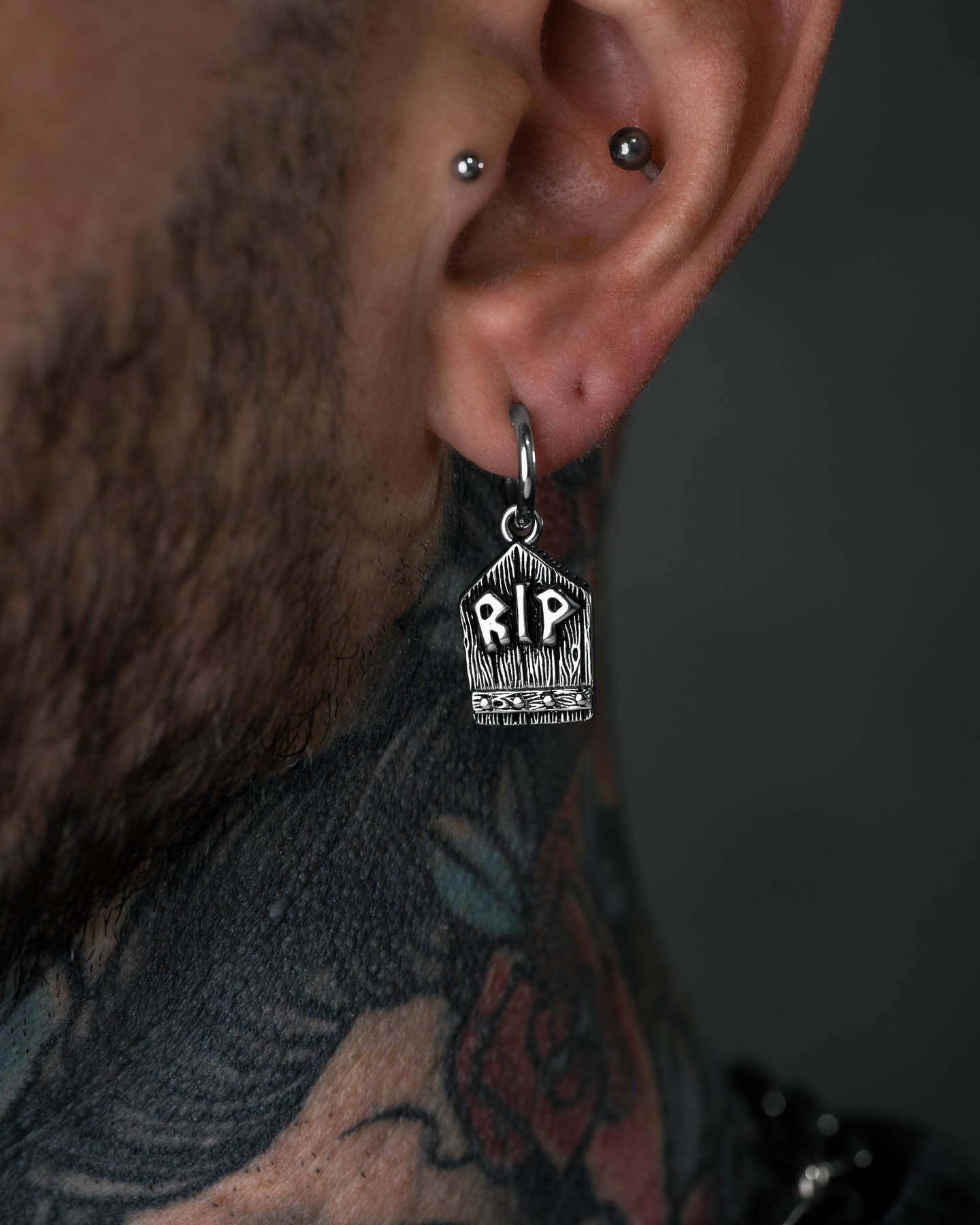 A close-up of a person's ear with two piercings, one wearing the Gravestone Earring engraved with RIP. Colorful tattoos on their neck enhance the unique and witchy vibe of this standout jewelry piece.