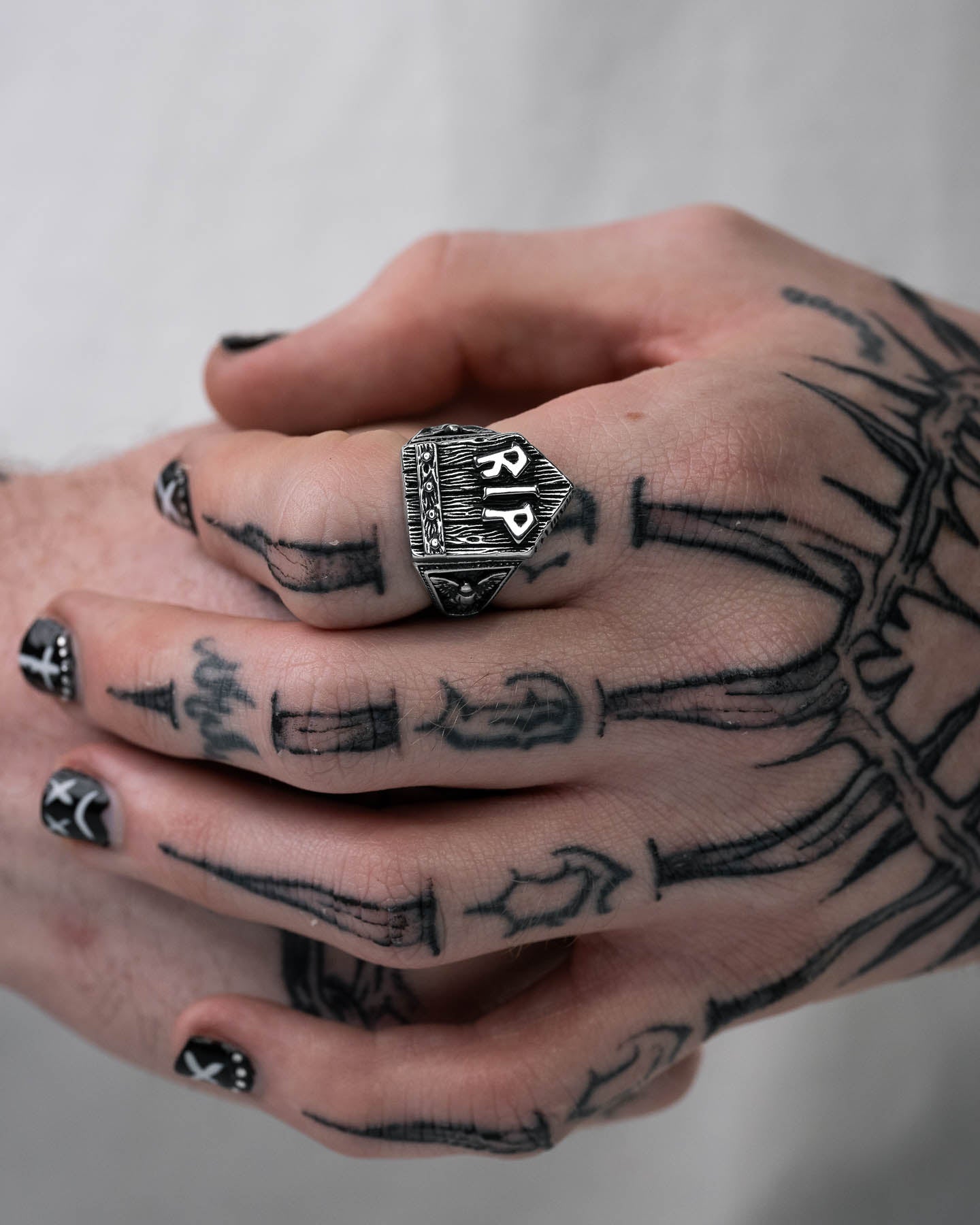 A close-up of tattooed hands with black and white bone designs. The person wears the Gravestone Ring, a large stainless steel ring engraved with RPG and geometric patterns. Their nails are painted black with white symbols.